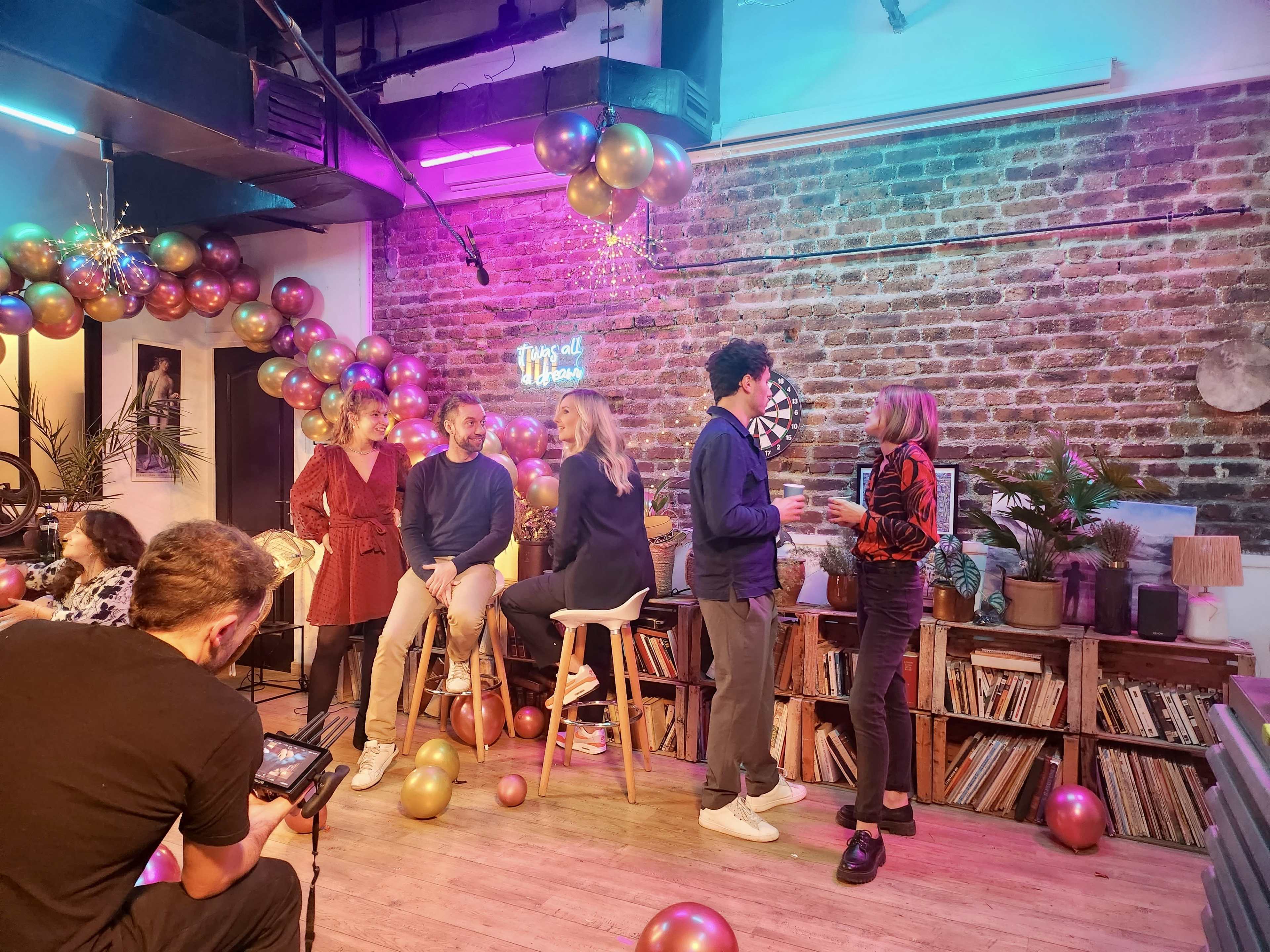 A group of six people chat and socialize in a dimly lit room adorned with balloons and string lights, while another individual records them.