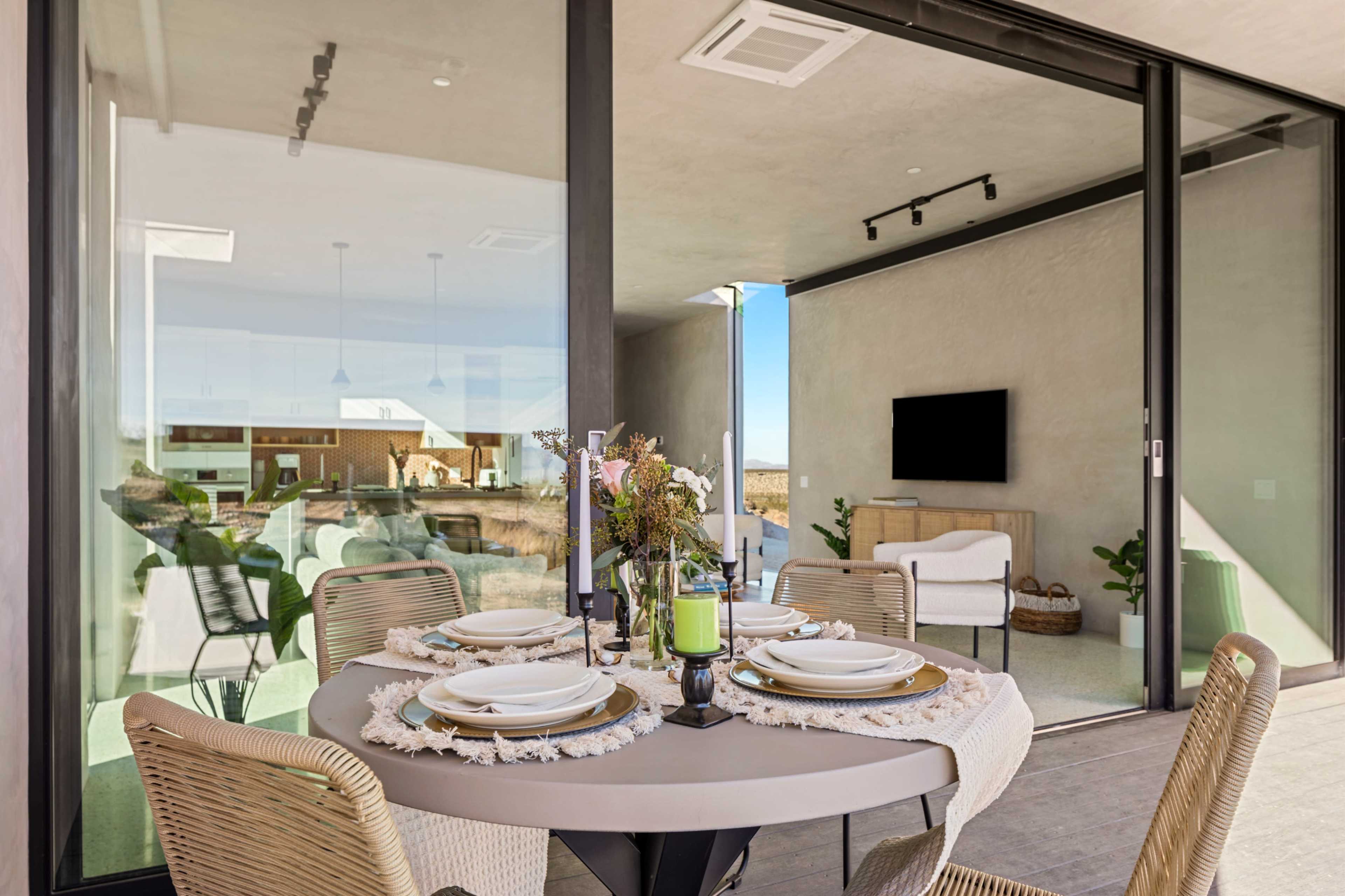 A modern dining area features a round table set with plates and a floral centerpiece, surrounded by large windows that open to a patio space.