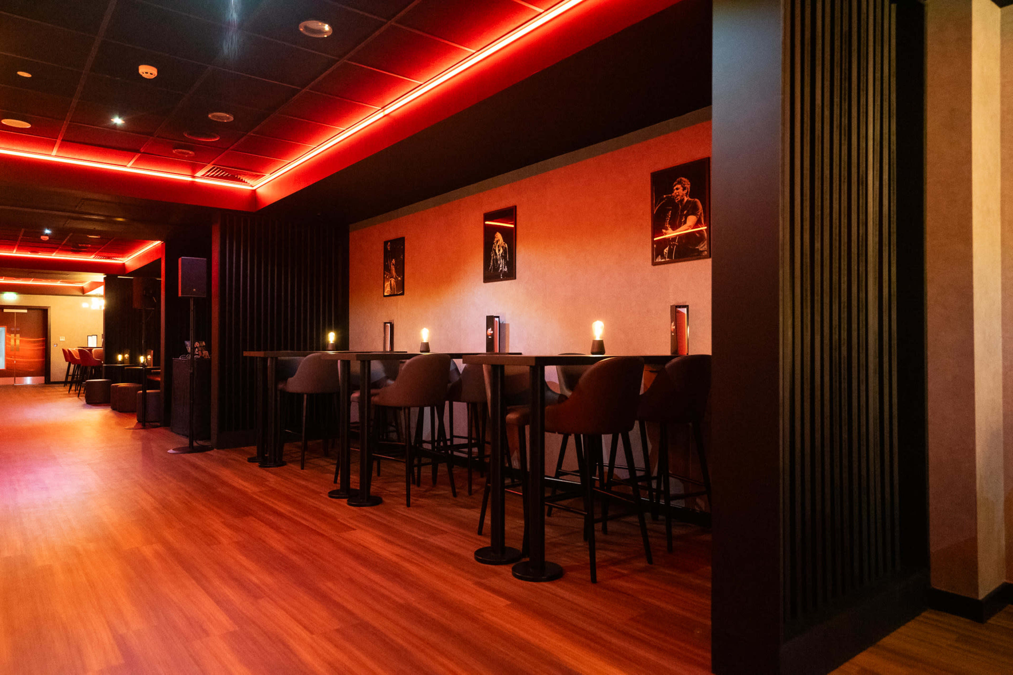 The image shows a modern bar area with tall tables and chairs, illuminated by red accent lighting and featuring framed photos on the walls.