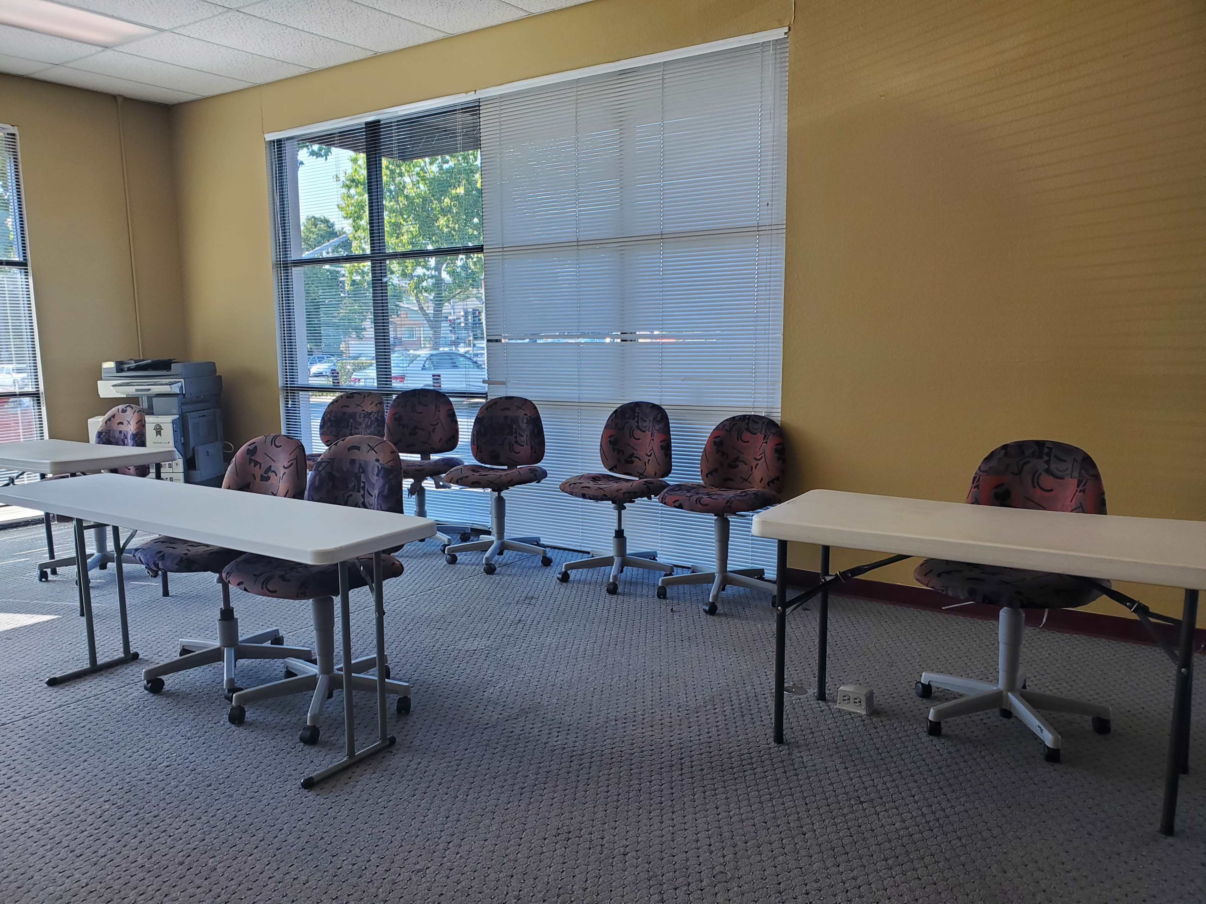 A room with two tables and several chairs arranged in a seating area, alongside windows letting in natural light.