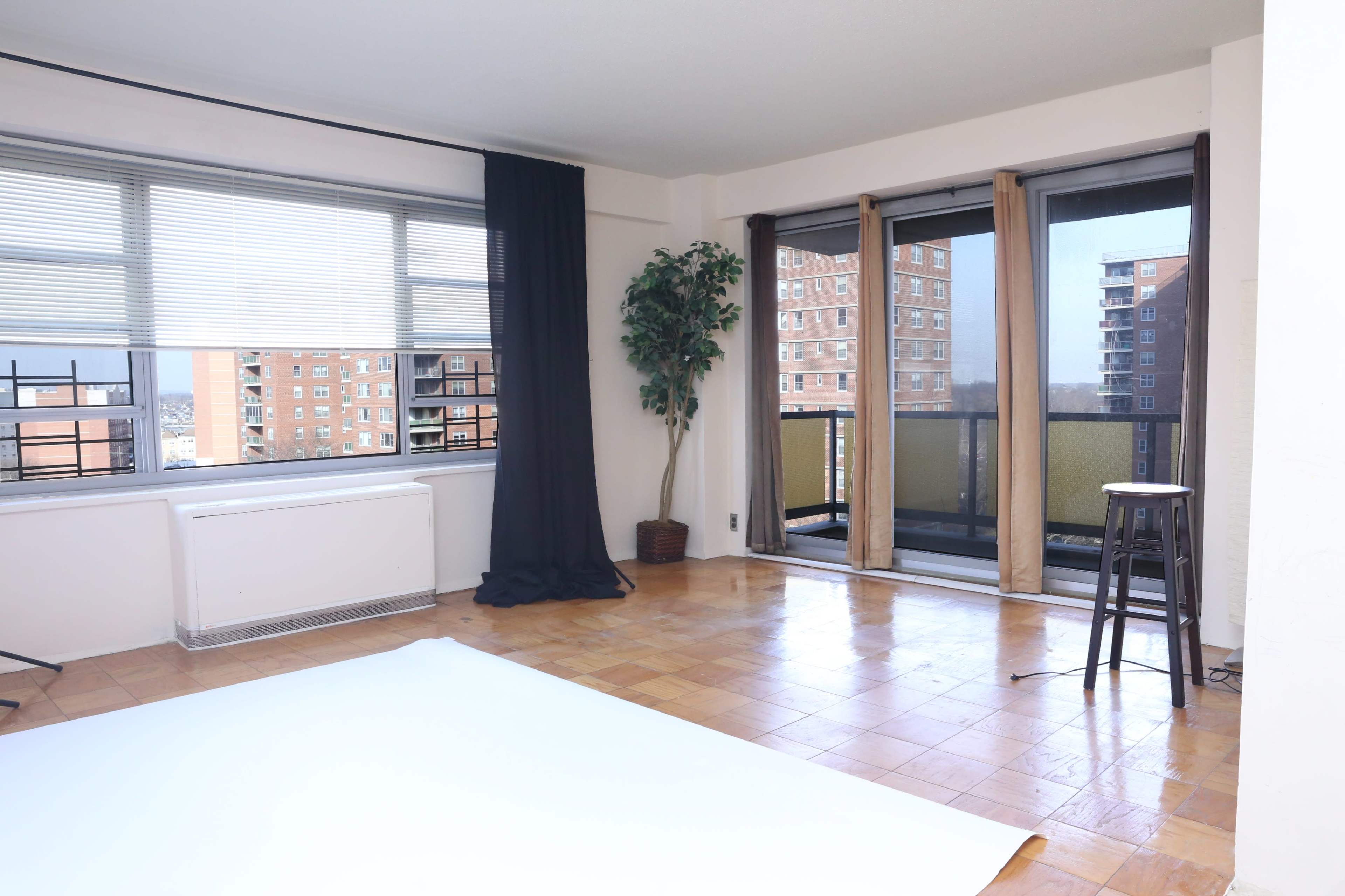 A spacious, empty room with large windows, wooden flooring, and a balcony, furnished with a potted plant and a stool.