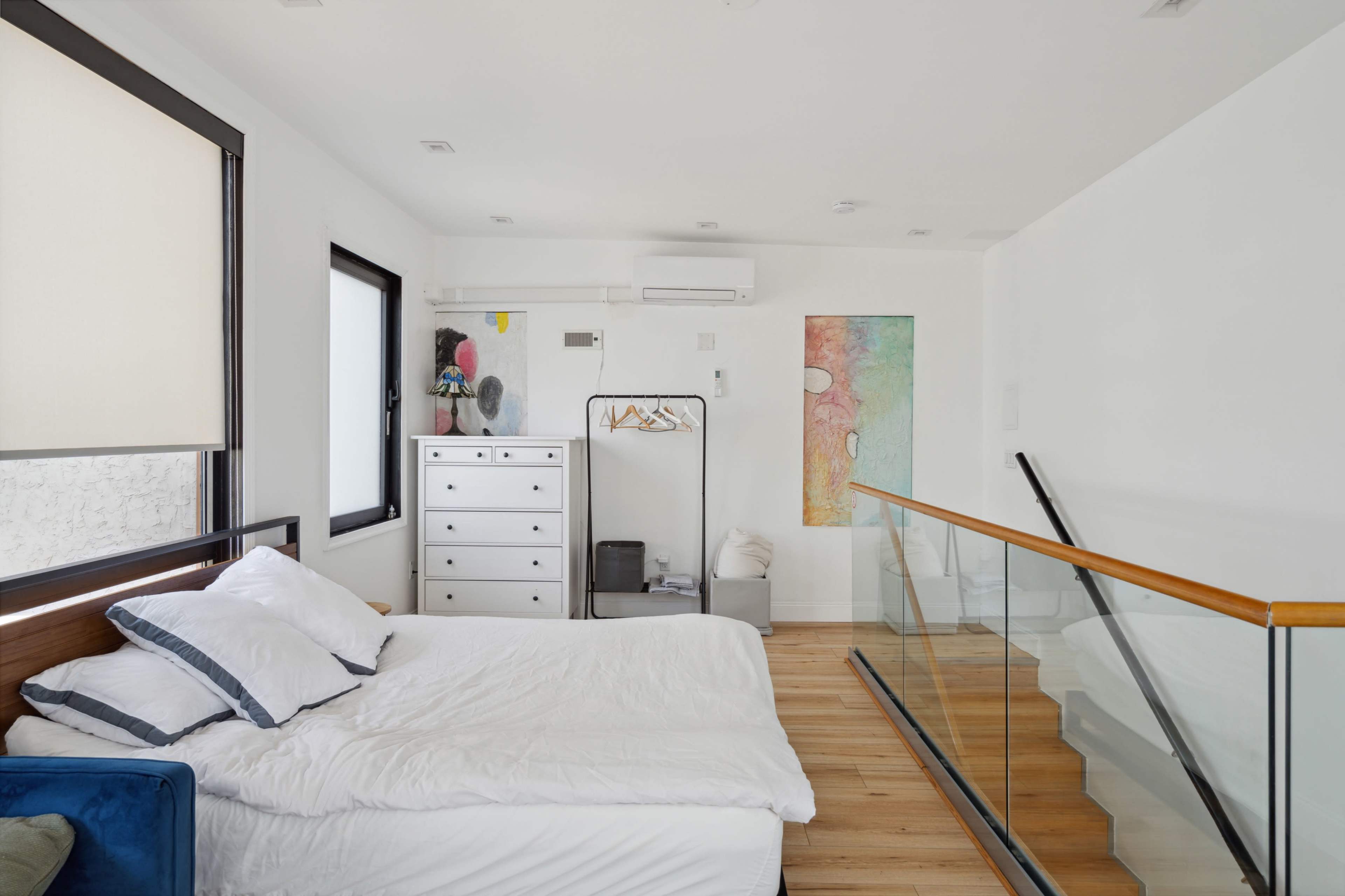 The image showcases a modern, minimalist bedroom featuring a unmade bed, a dresser, and a clothing rack, with large windows allowing natural light to enter the space.