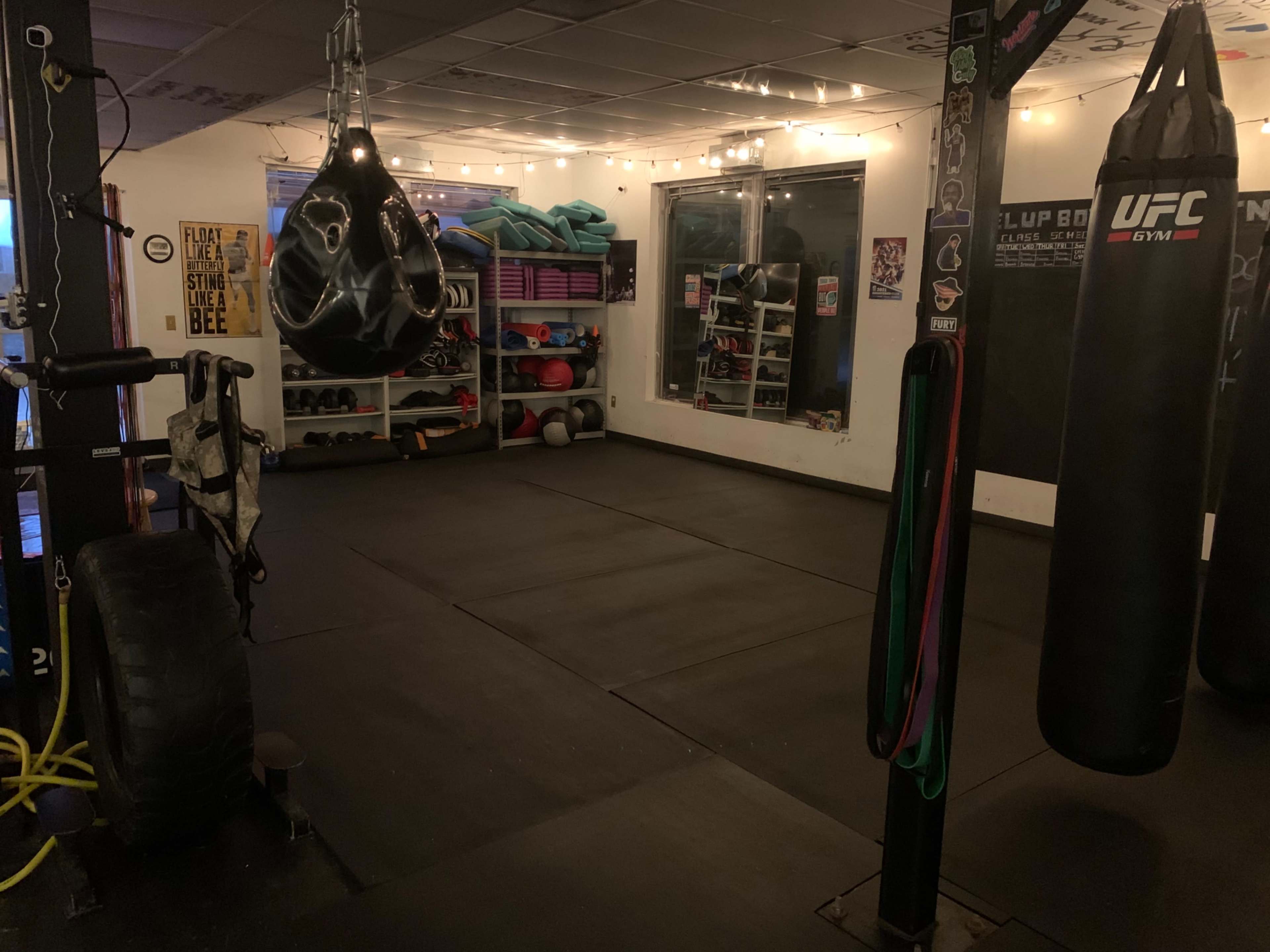 A dimly lit gym space with heavy bags, exercise equipment, and a rubber mat flooring, along with shelves for storage.