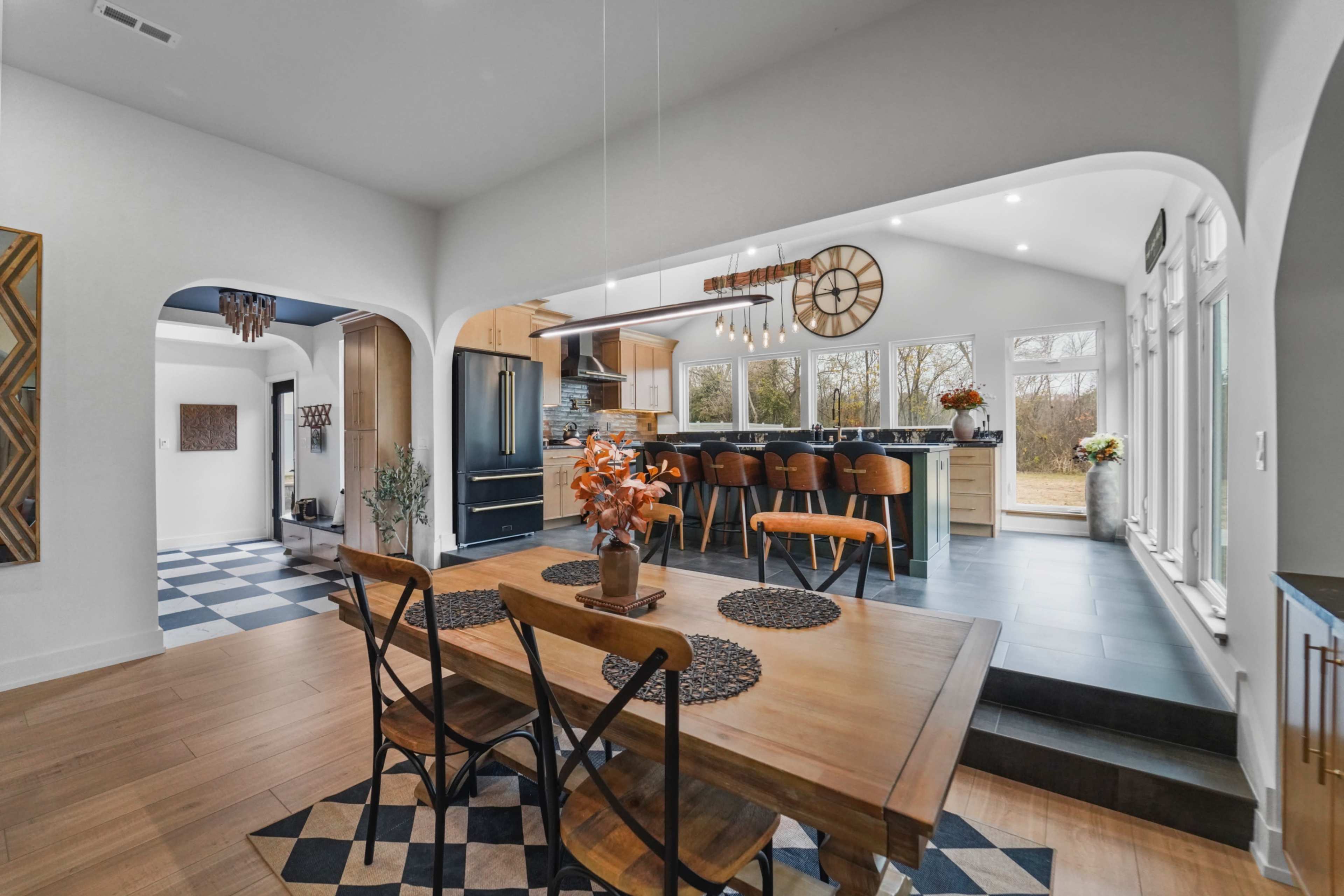 The image shows an open-concept kitchen and dining area with a wooden dining table, modern kitchen appliances, and large windows providing natural light.