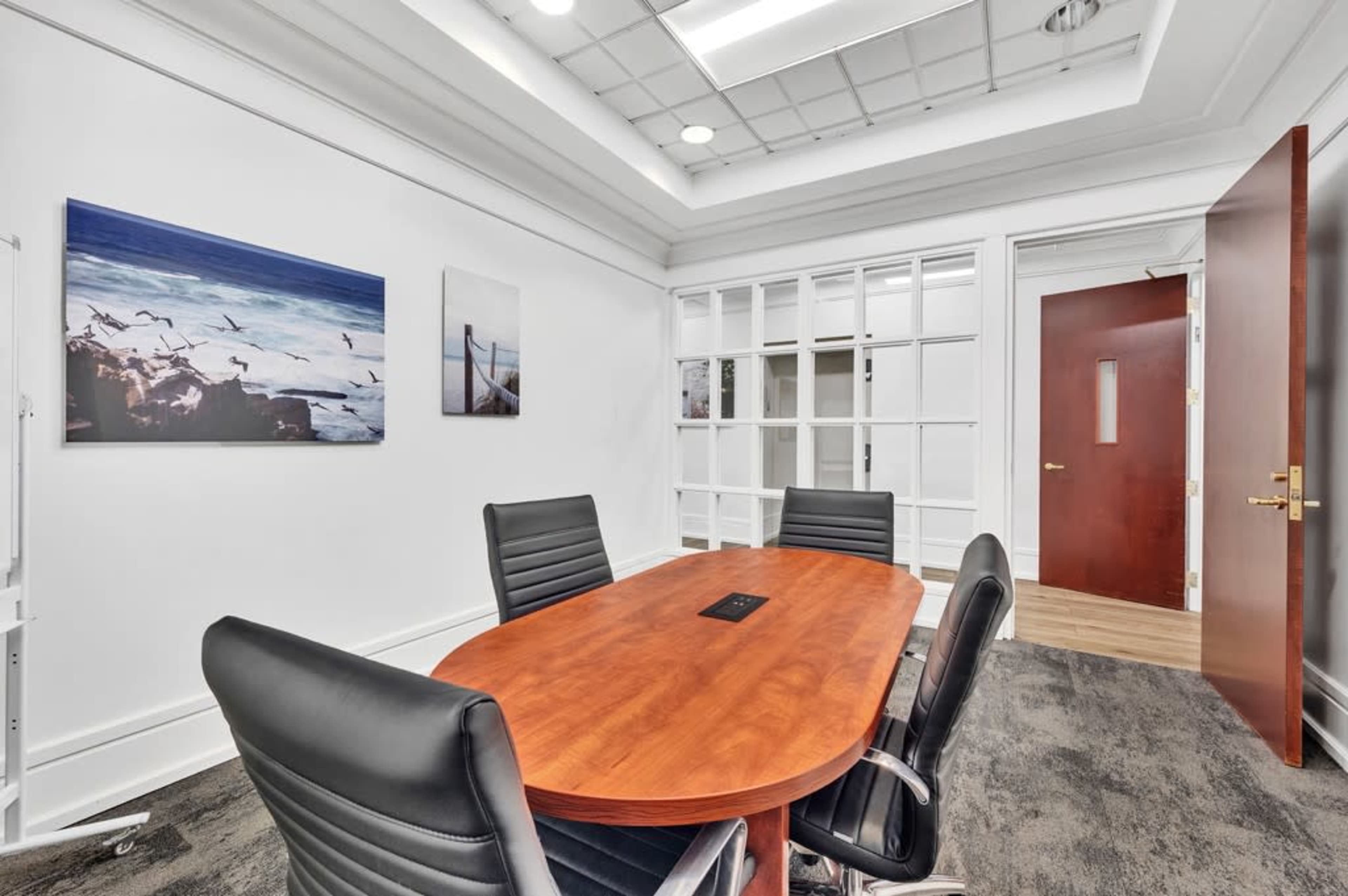 The image shows a conference room featuring a wooden oval table surrounded by black chairs, with framed pictures on the walls and a glass partition leading to another area.