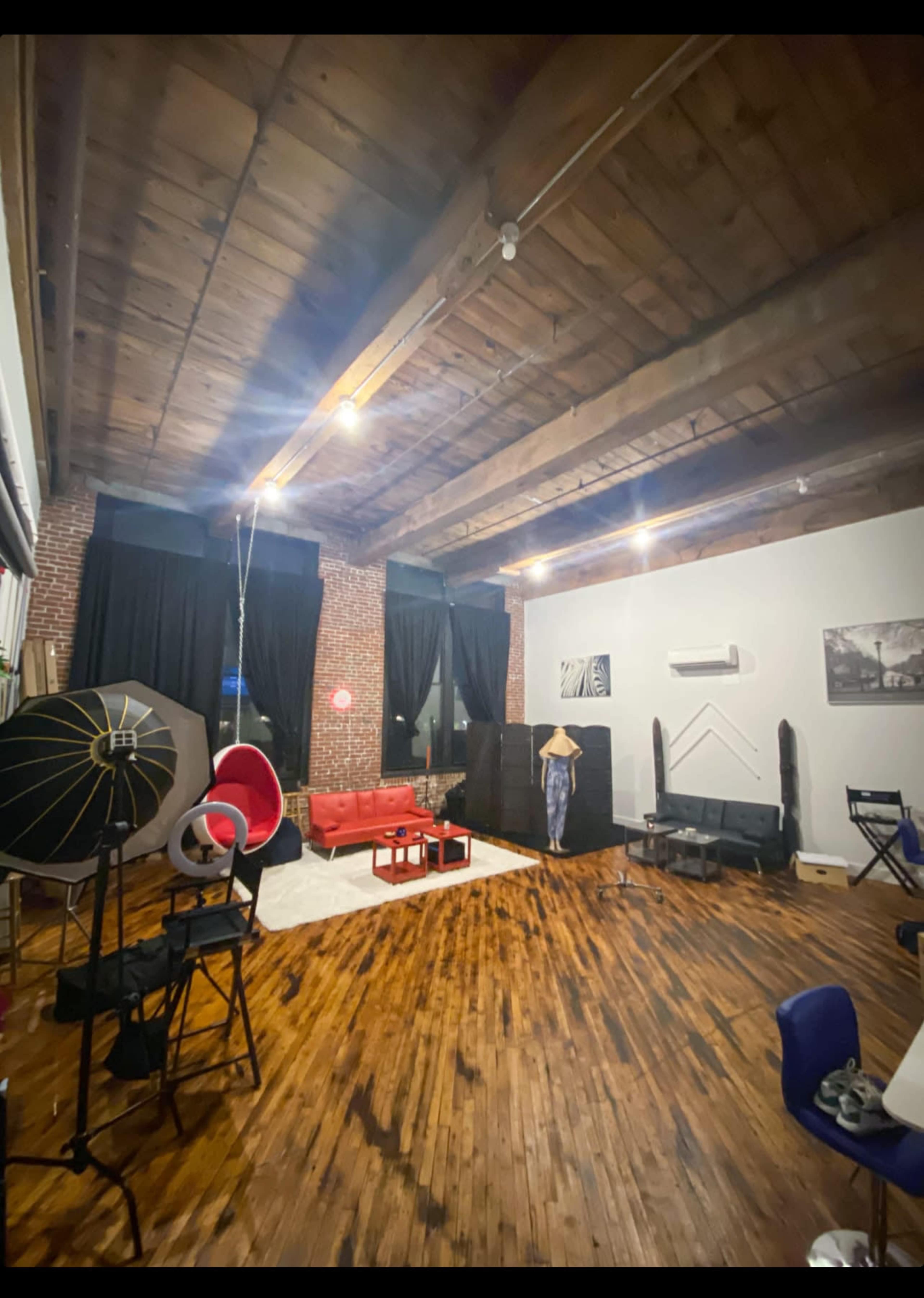 A spacious, well-lit loft featuring wooden floors, a red sofa, various chairs, and photography equipment against exposed brick walls.