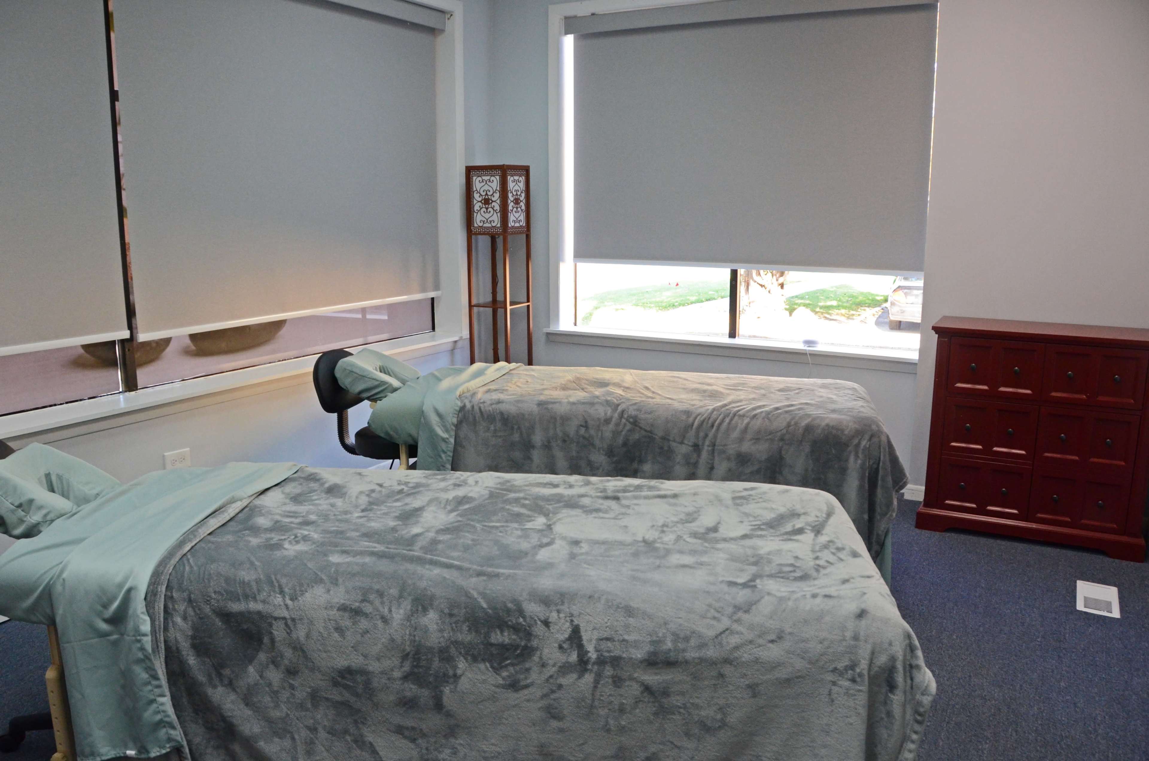 A massage room with two massage tables covered in grey linens, a chair, and a red wooden cabinet.