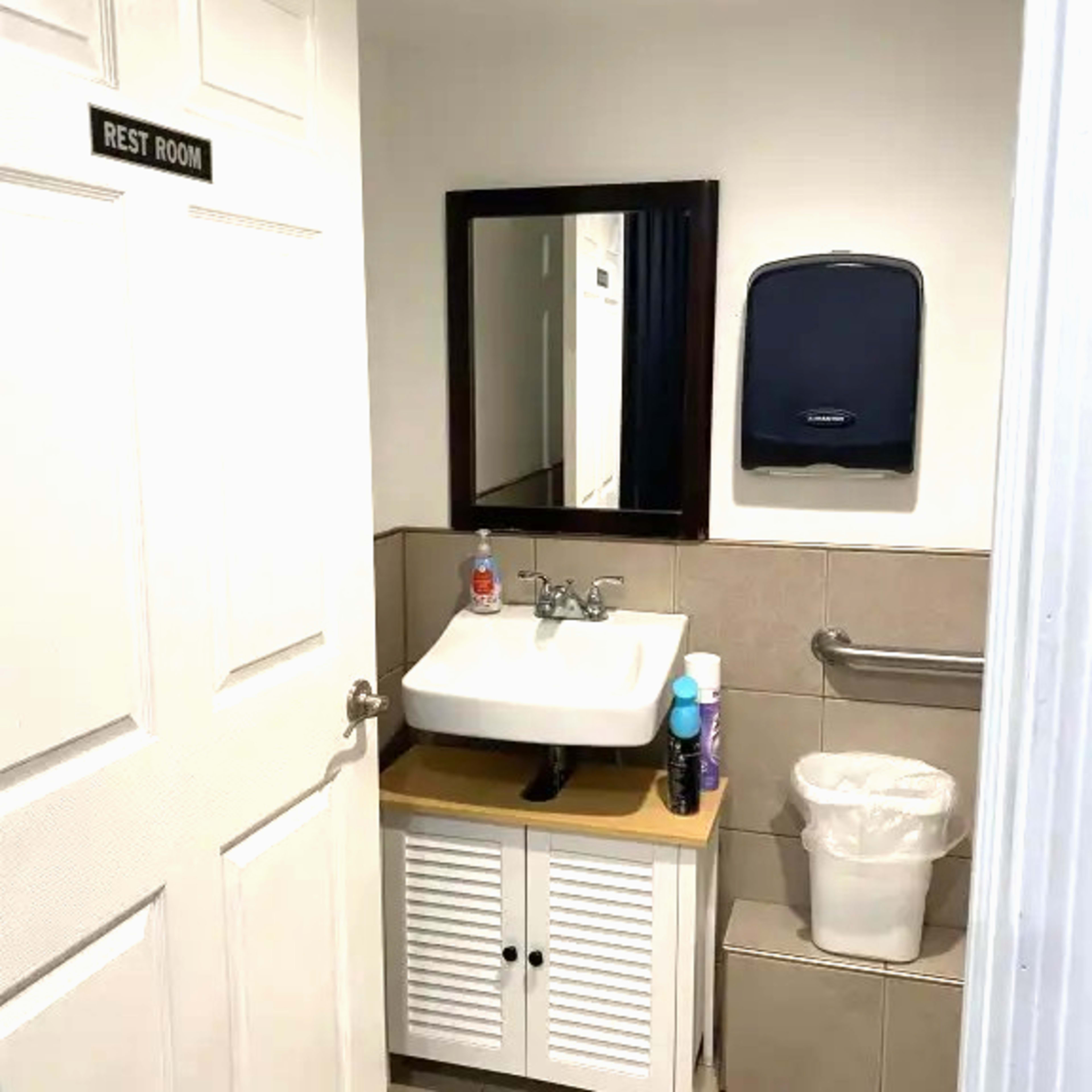 The image shows a small restroom with a sink, a mirror above it, hand sanitation supplies, and a wastebasket, all situated beside a closed door labeled "REST ROOM."