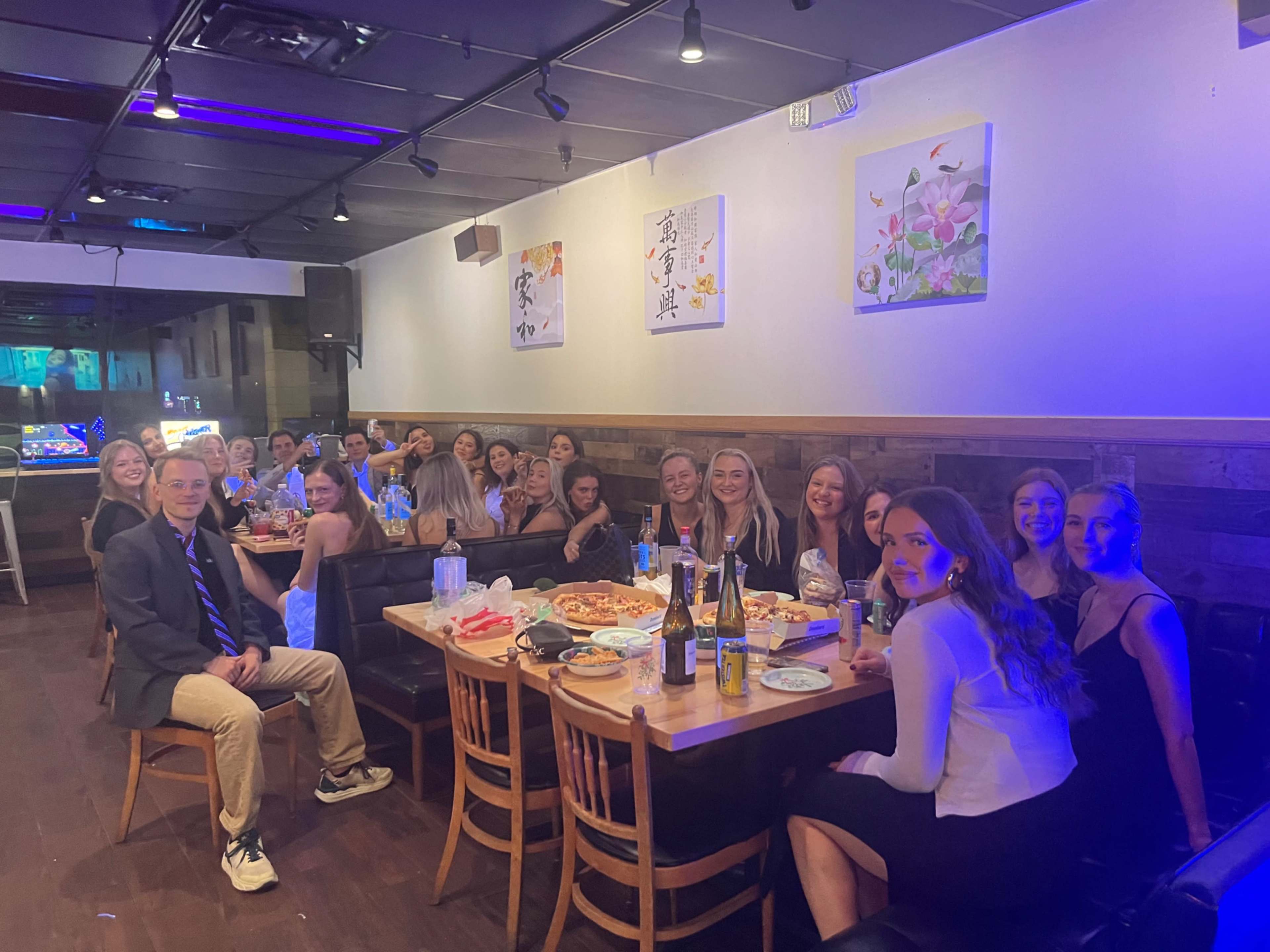 A large group of young people sits at tables in a restaurant, enjoying food and drinks, with colorful lighting and decorative artwork on the walls.