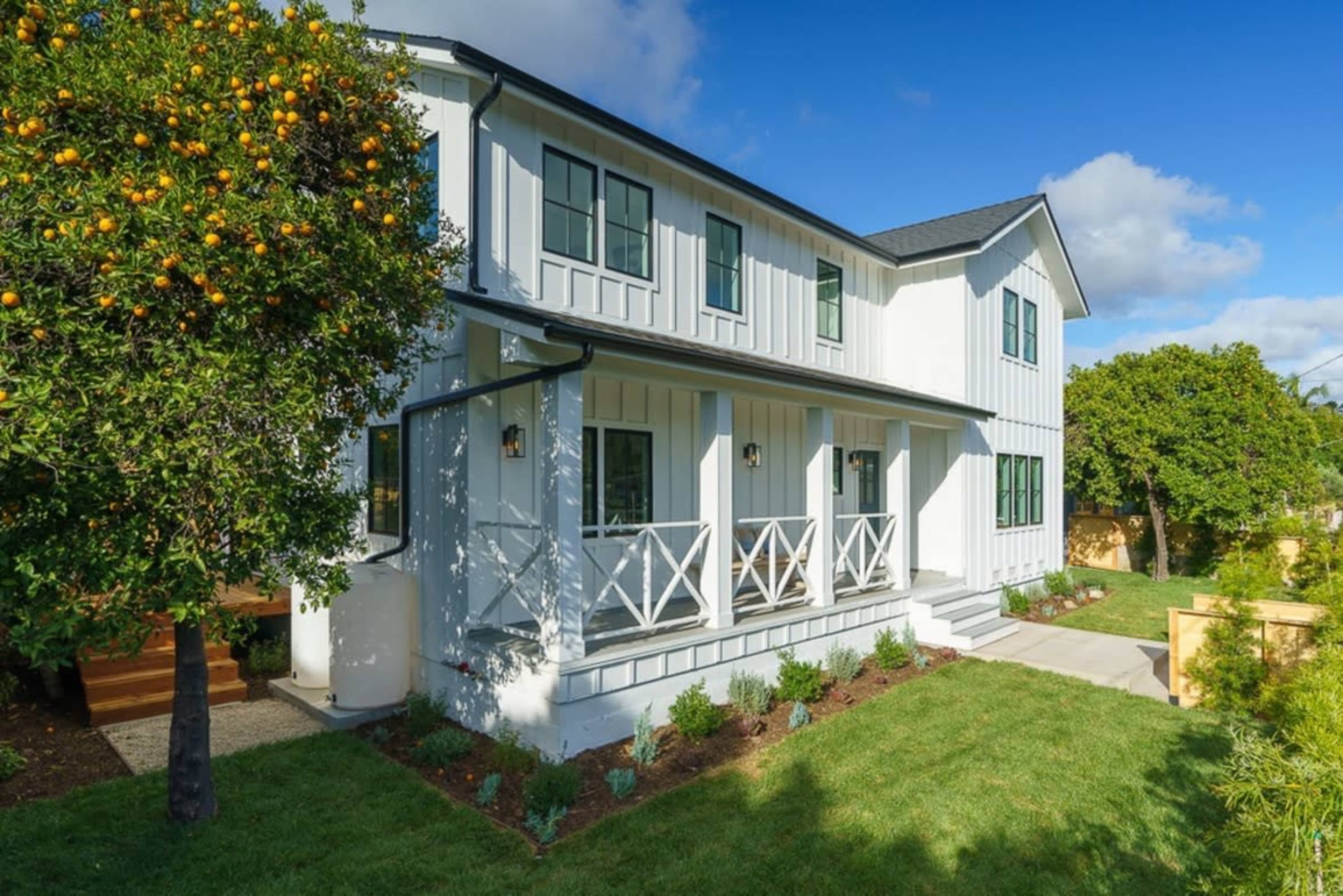 A modern two-story white wooden house with a porch and a landscaped yard featuring a citrus tree.