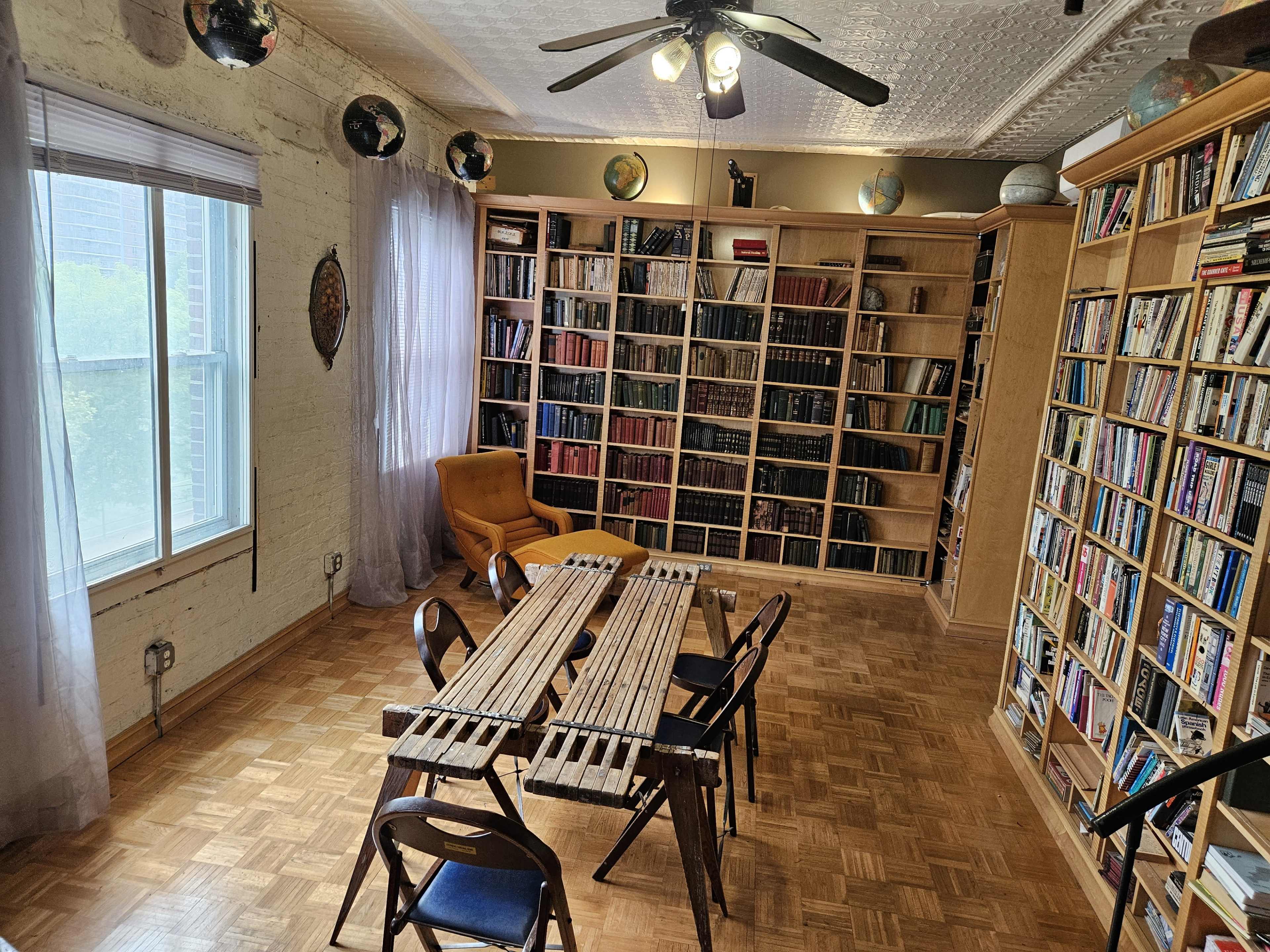 The image shows a cozy library with wooden bookshelves filled with books, a round table made of slats, and a bright yellow chair beside a large window.