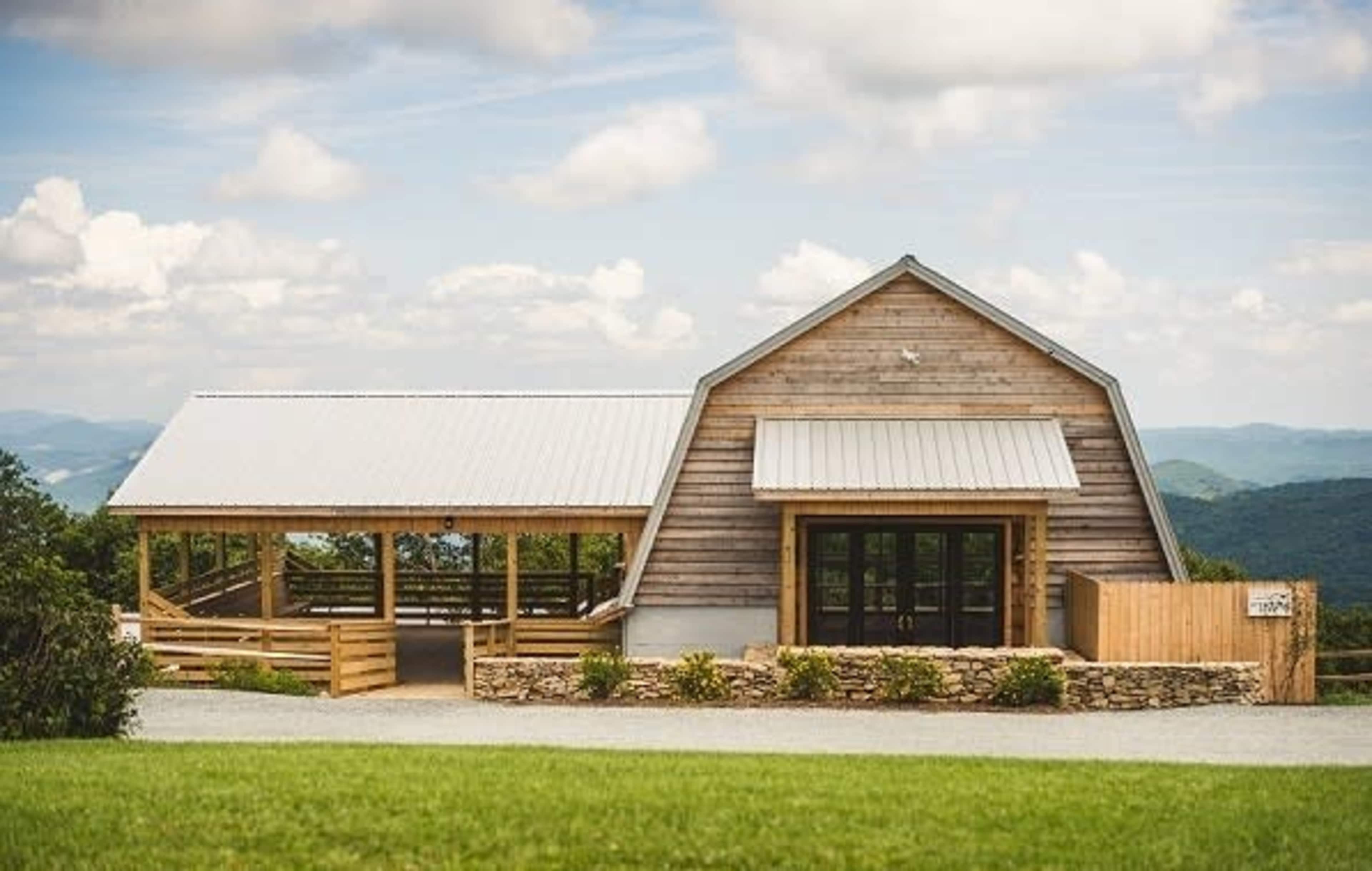 A wooden structure with a gabled roof, featuring large glass doors and a covered porch, situated on a grassy area with mountains in the background.