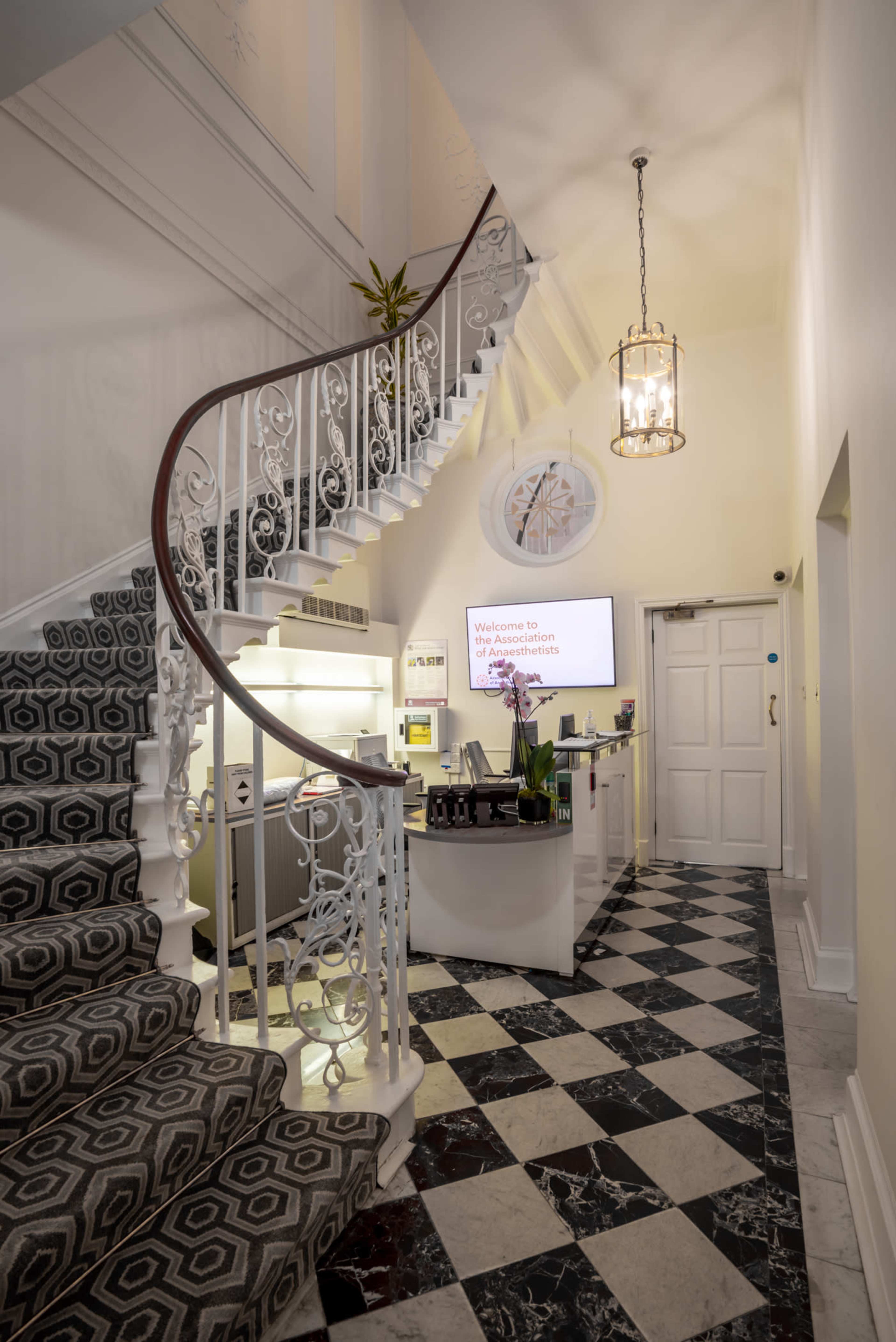 A well-lit reception area featuring a curved staircase with decorative railings, a modern front desk, and a patterned tiled floor.