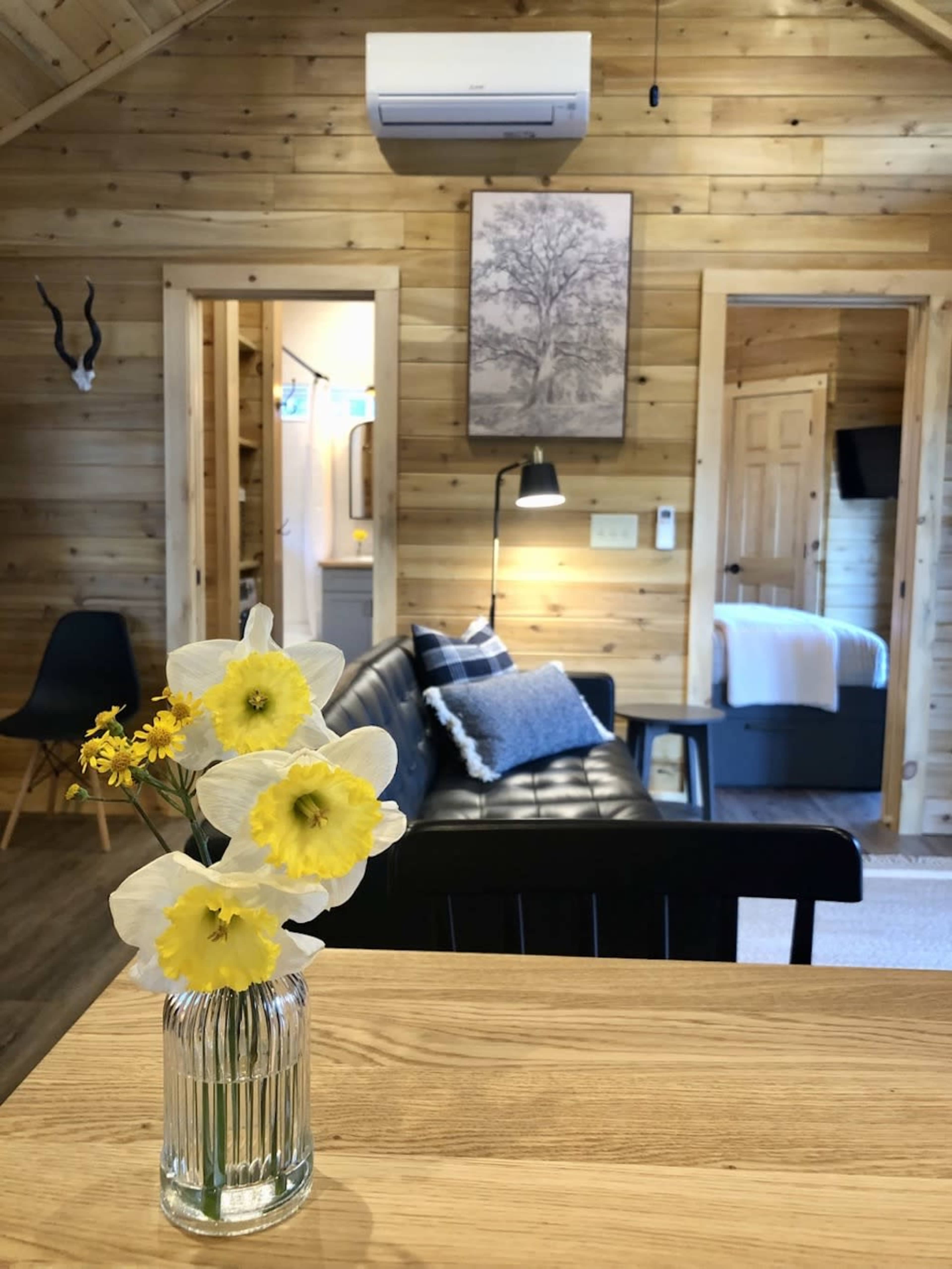 A vase with yellow flowers sits on a wooden table in a cozy, rustic interior featuring a leather couch and wooden walls.
