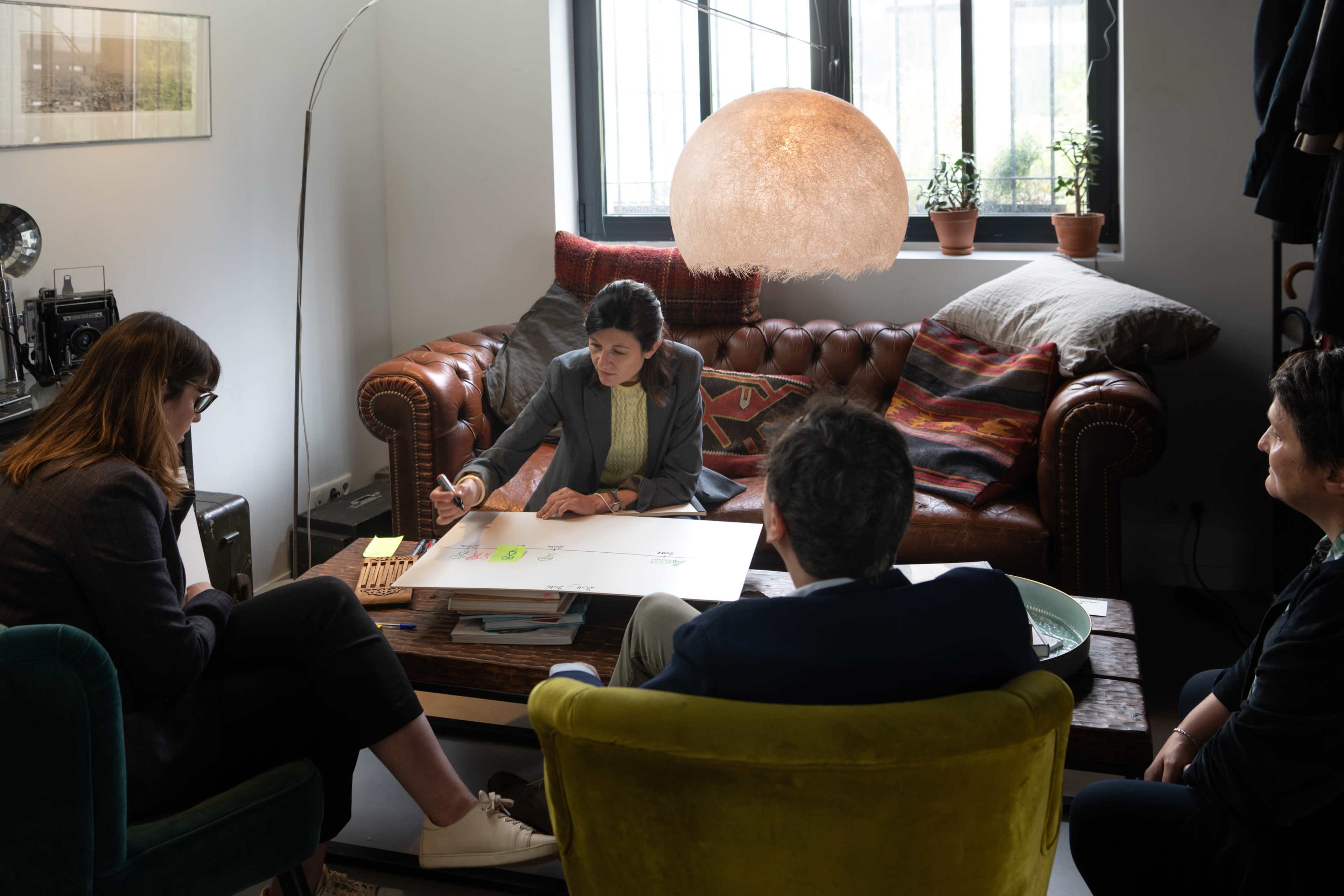 A group of four people is engaged in a discussion around a table with a large sheet of paper, with one person writing on it while sitting on a sofa in a cozy, well-lit room.