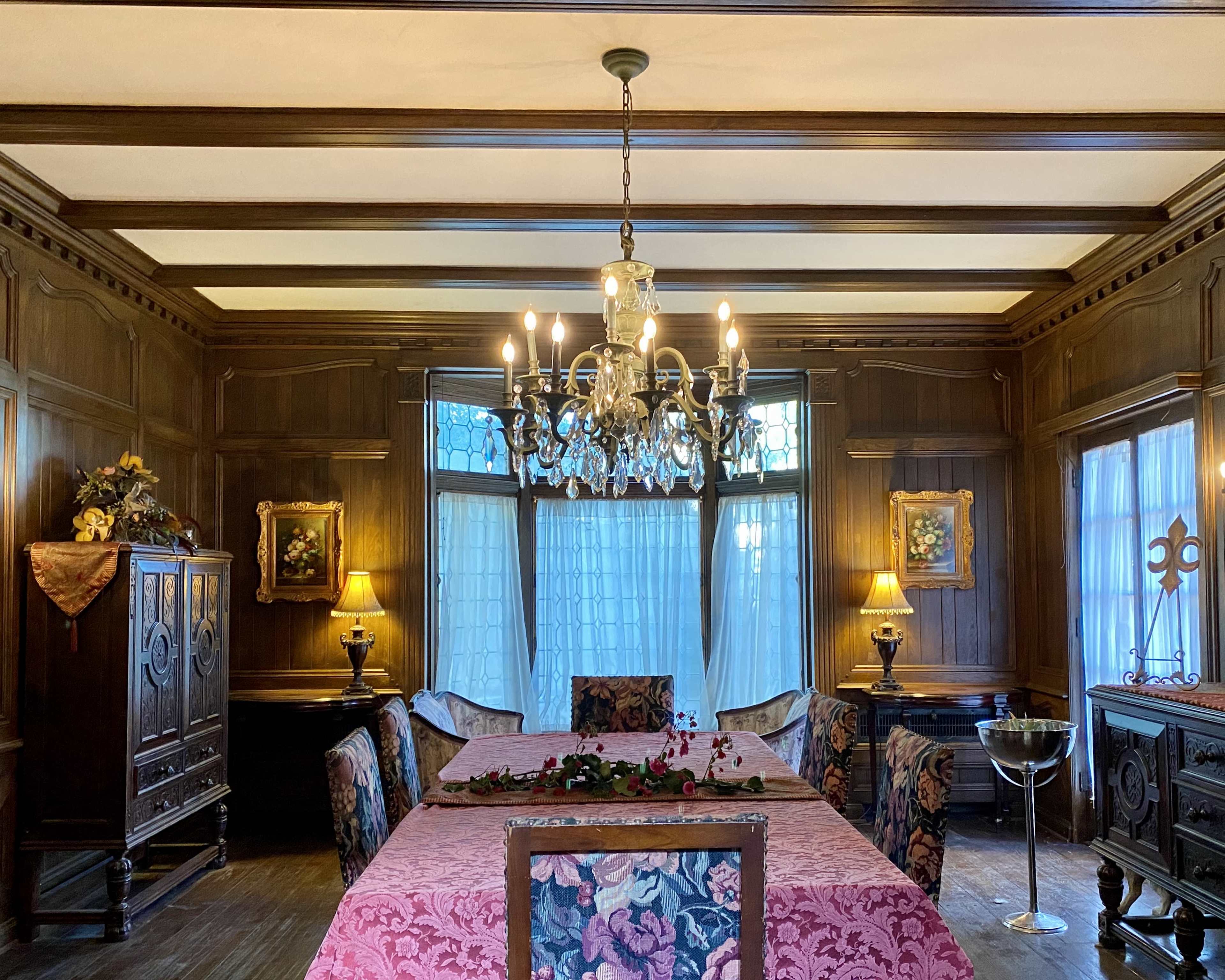 An elegantly furnished dining room featuring a long table with a pink tablecloth, surrounded by upholstered chairs, and illuminated by a crystal chandelier.