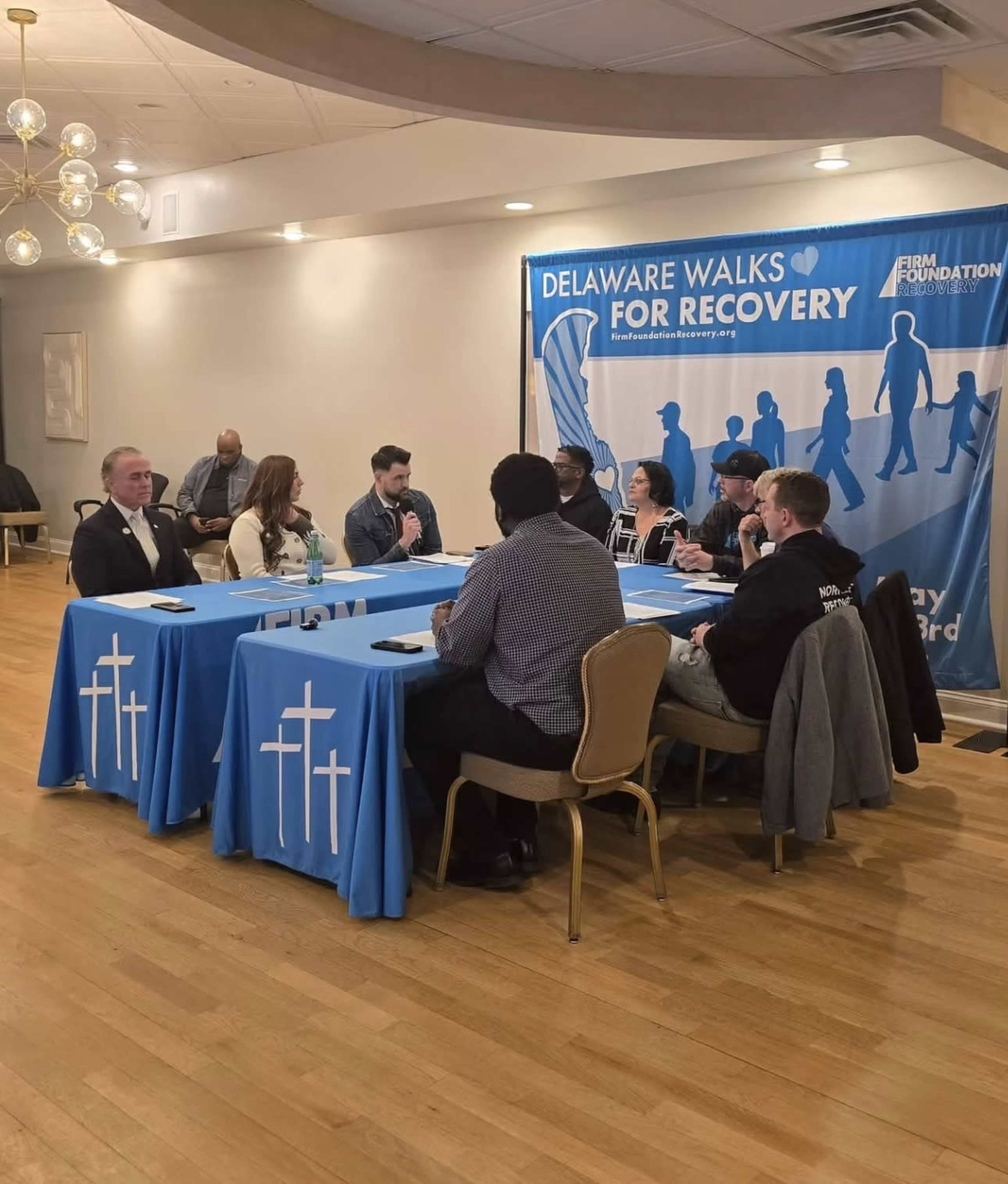 A group of people is seated around a table with a blue tablecloth displaying white crosses, discussing topics related to recovery in front of a banner that reads "Delaware Walks for Recovery."