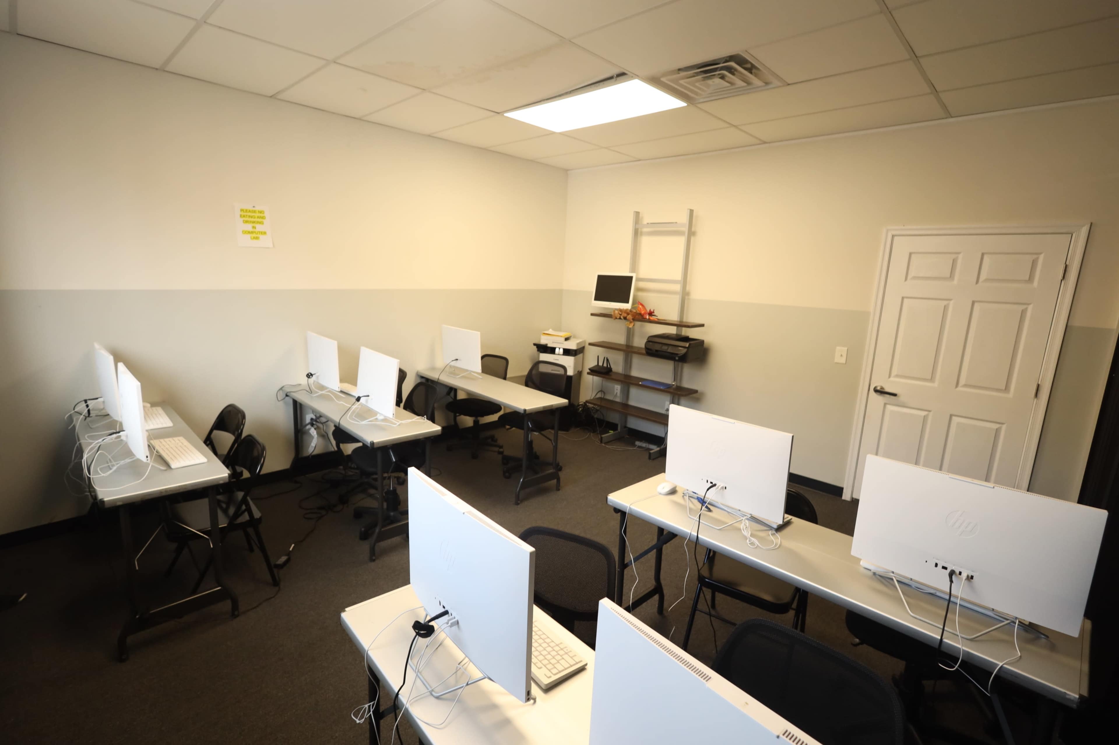 The image shows a classroom with several computer stations arranged in rows, featuring desks and chairs, along with a wall-mounted shelf containing a printer.