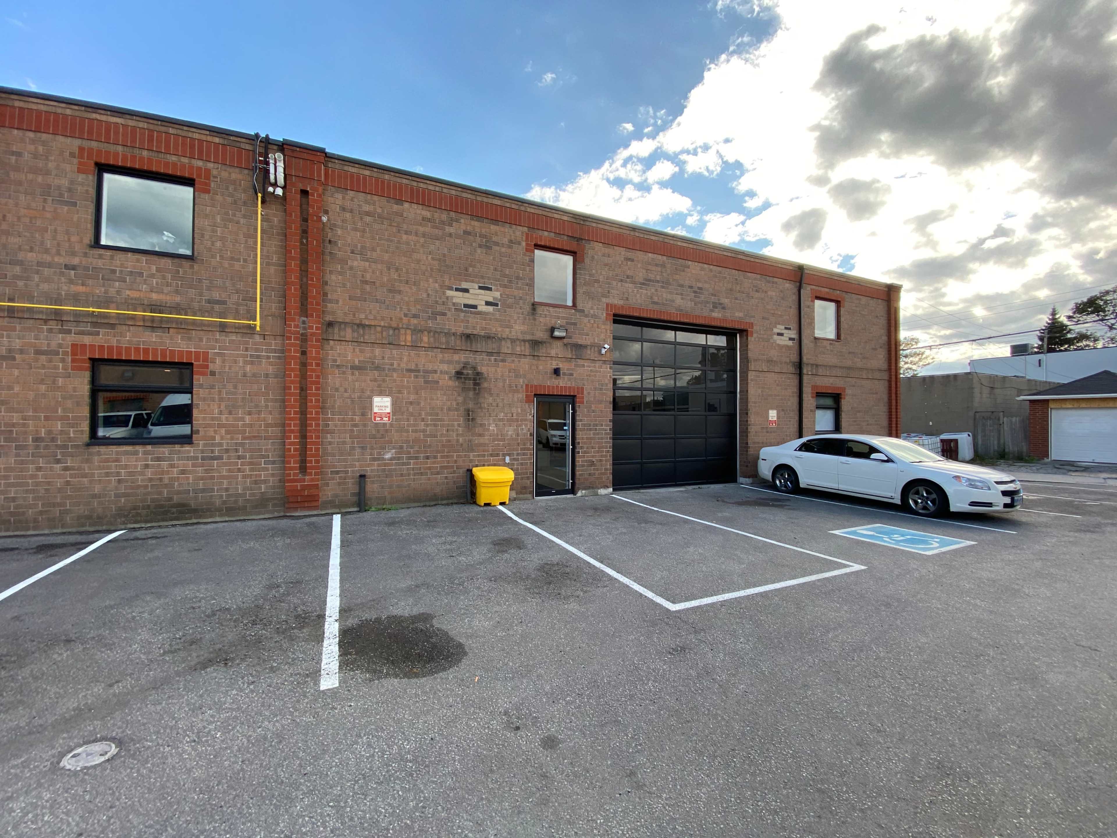 A brick commercial building with a large garage door, a parked white car, and a yellow waste bin in the parking lot.