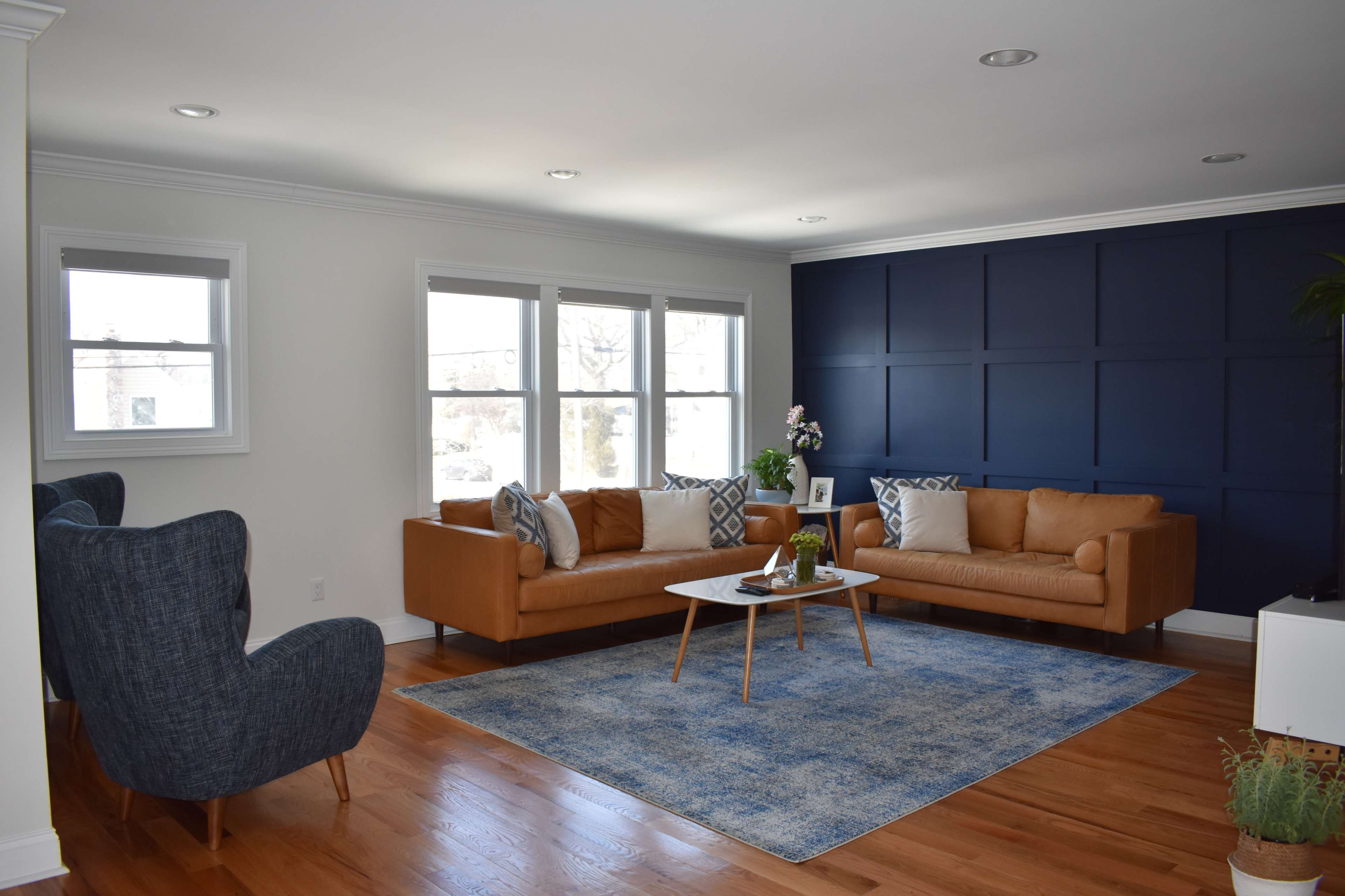 A contemporary living room with a brown leather sofa, a blue area rug, and a dark blue accent wall, complemented by a coffee table and two upholstered chairs.