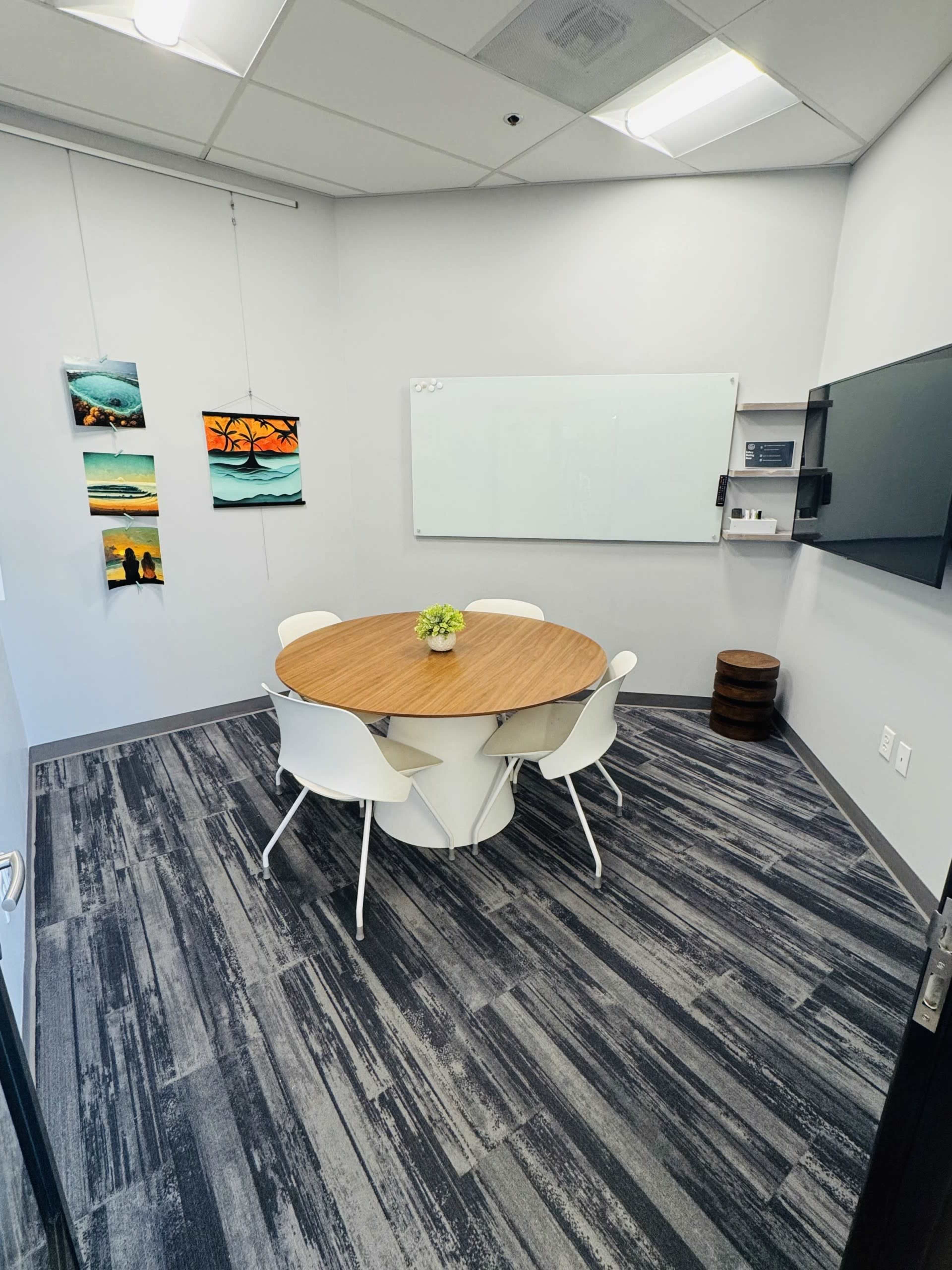 The image shows a small conference room with a round wooden table surrounded by four white chairs, two pieces of artwork on the wall, a whiteboard, and a mounted screen.