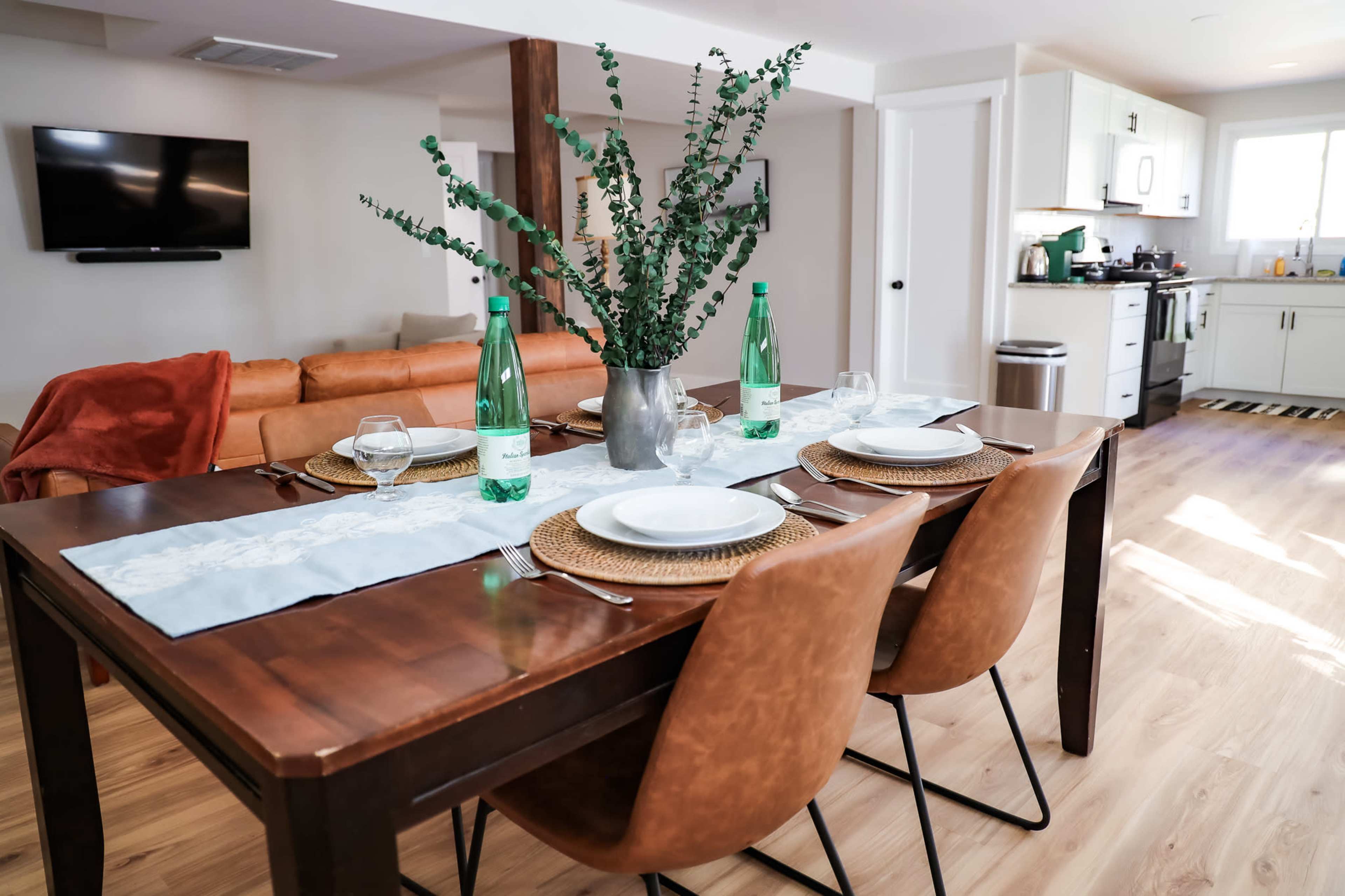 A dining table is set for a meal with glassware and plates, accompanied by a vase of greenery, in a bright and open kitchen-living space featuring a couch and a television.
