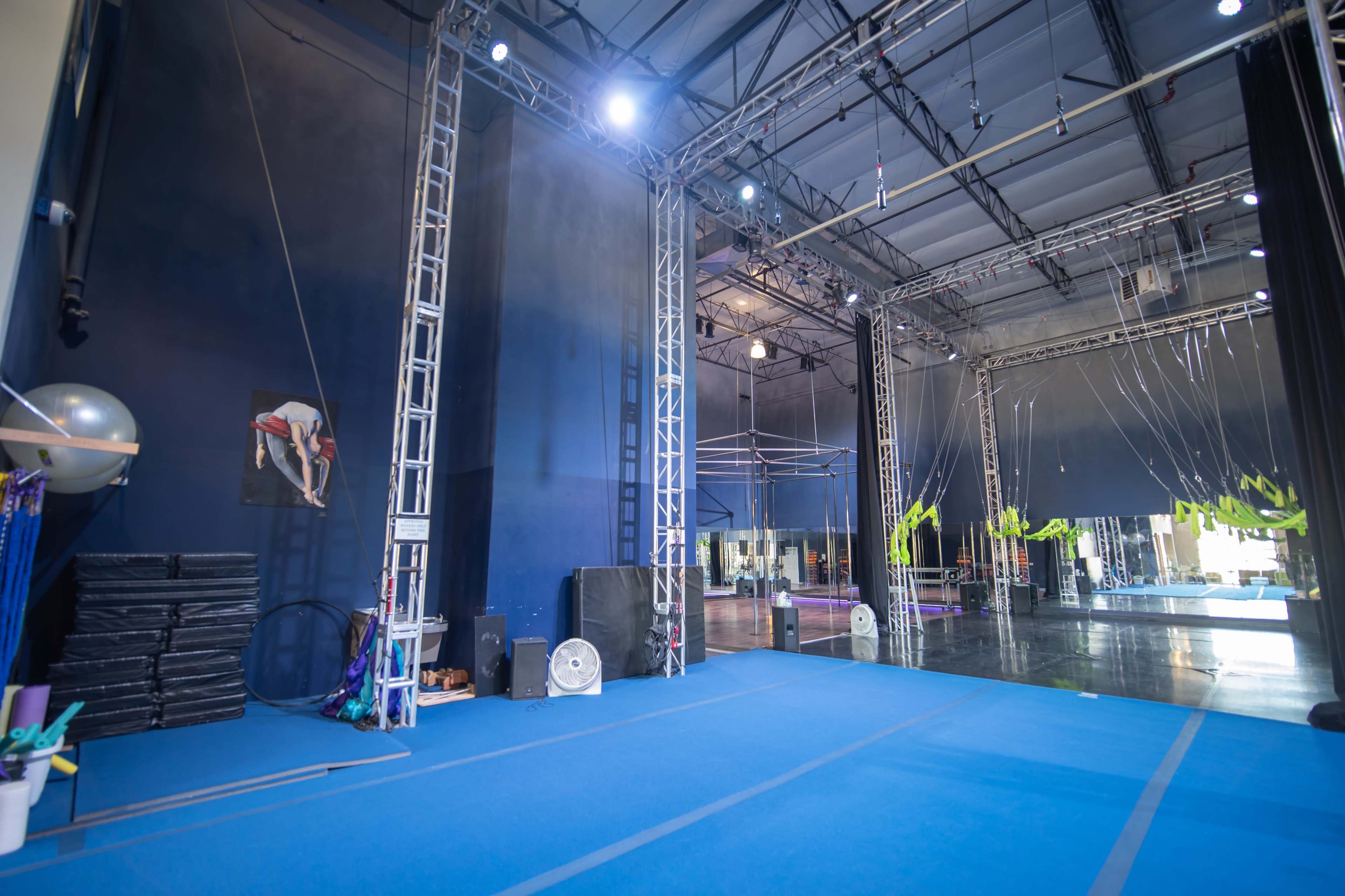 An indoor gym space with blue flooring, framed by metal trusses and equipment, including a suspended aerial apparatus and storage mats.