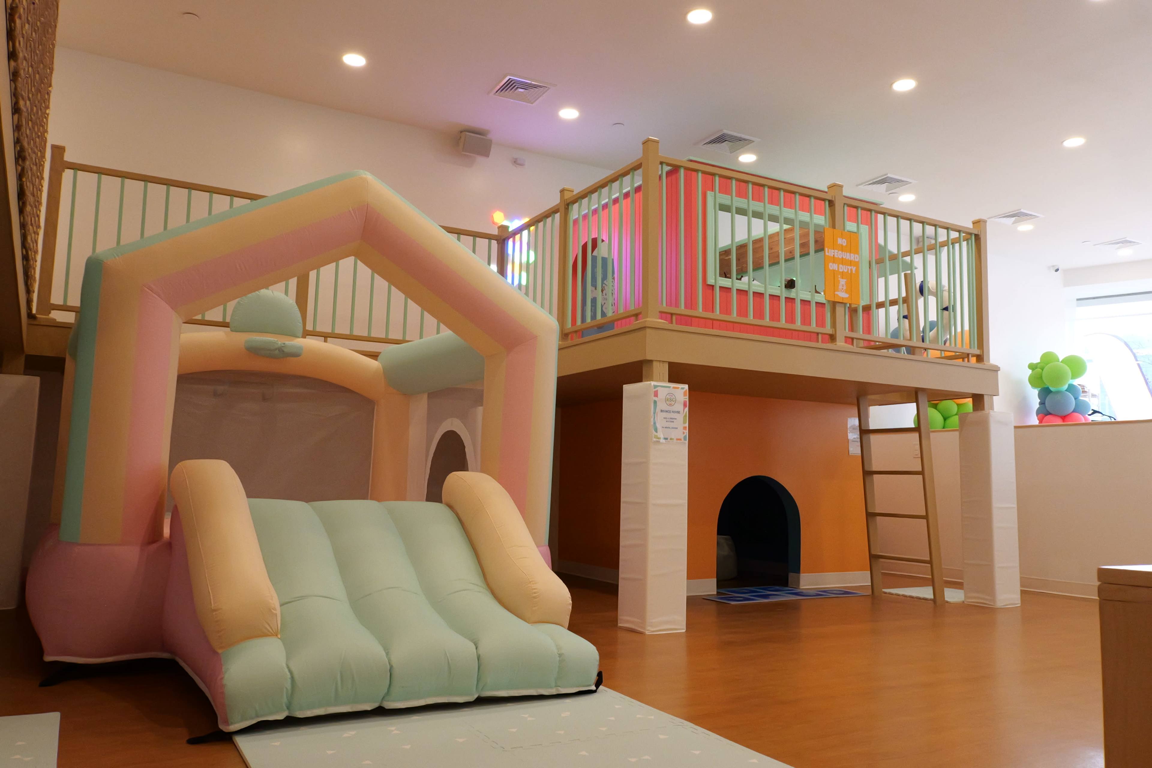The image shows a colorful indoor play area featuring a bounce house with a slide and a wooden play structure with a ladder.