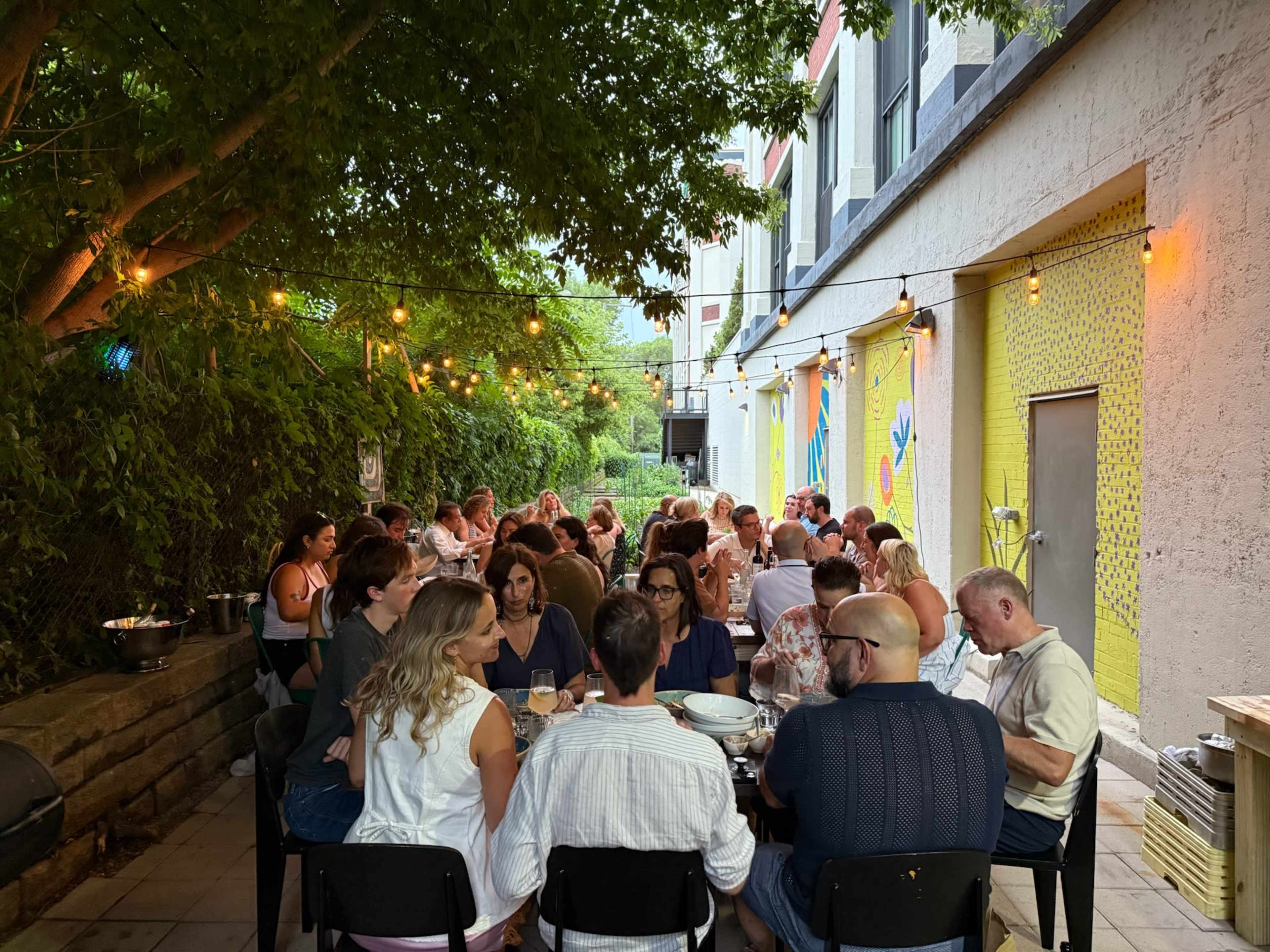 A large group of people is seated at long tables outdoors under string lights, with a mural on a yellow wall in the background.
