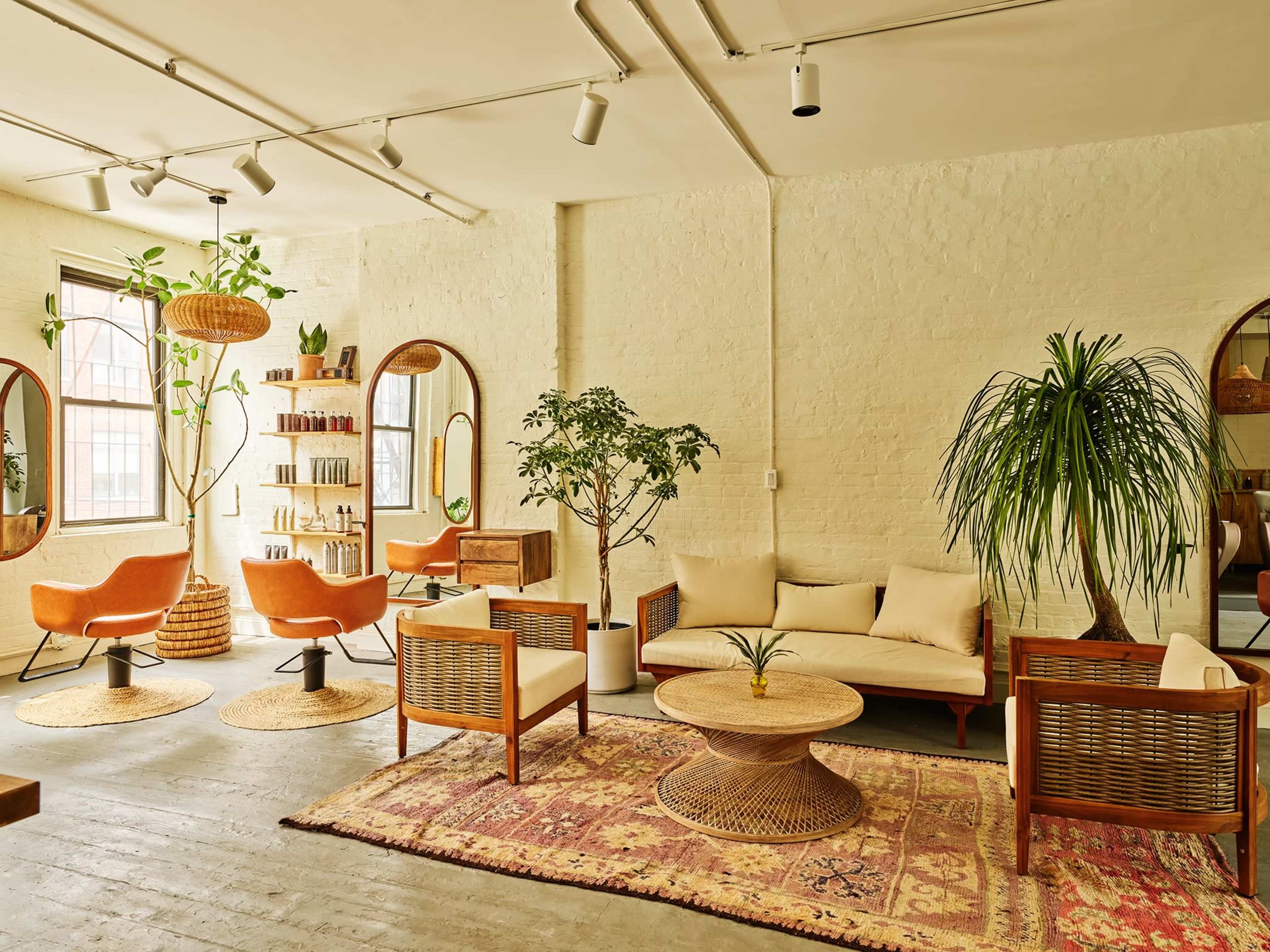 The image shows a stylish interior of a salon featuring orange chairs, a large round coffee table, and plants against a light-colored wall.