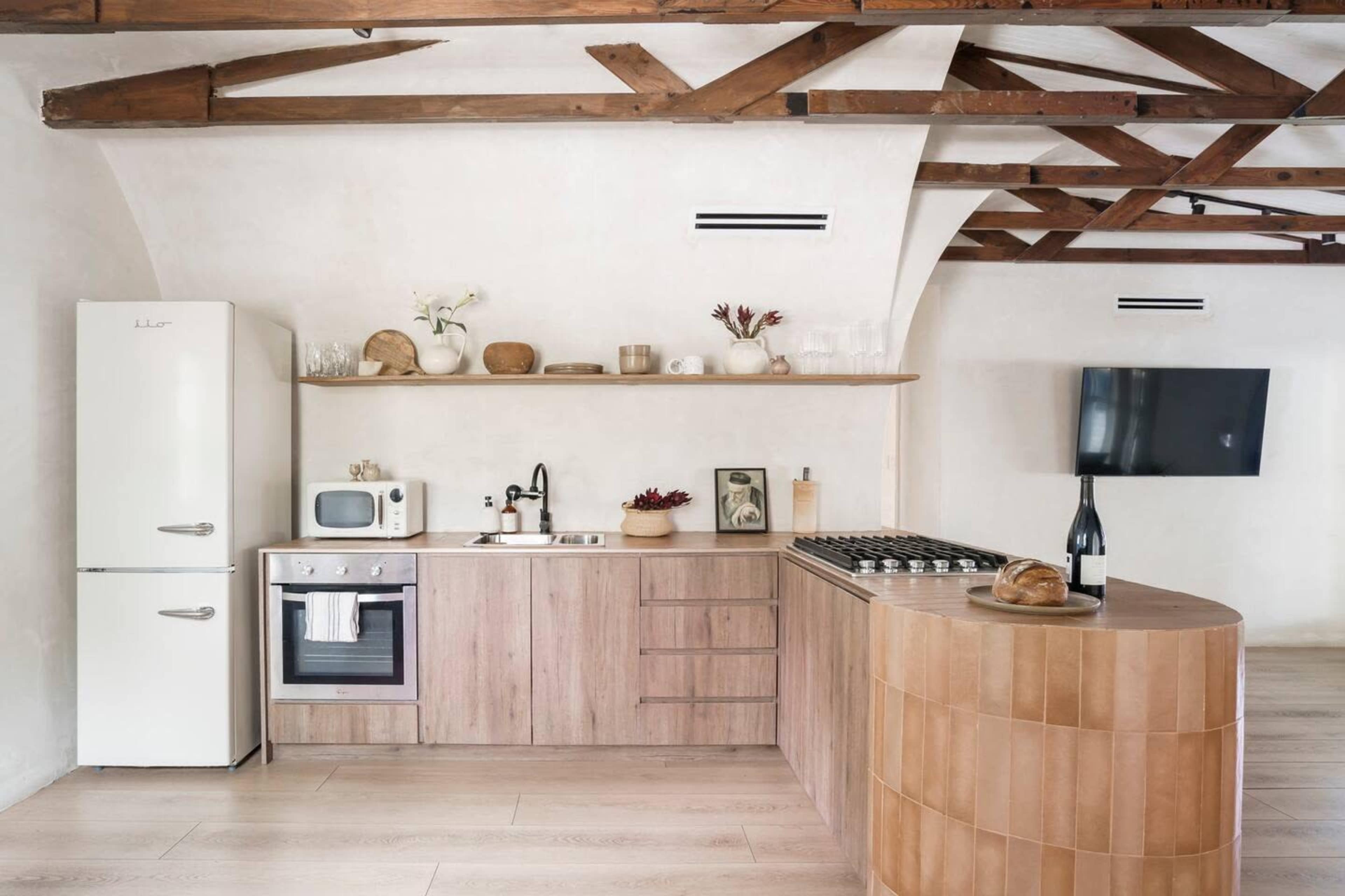 The image shows a modern kitchen with wooden cabinetry, a white refrigerator, and a curved wooden island, set against a backdrop of exposed beams and light-colored walls.