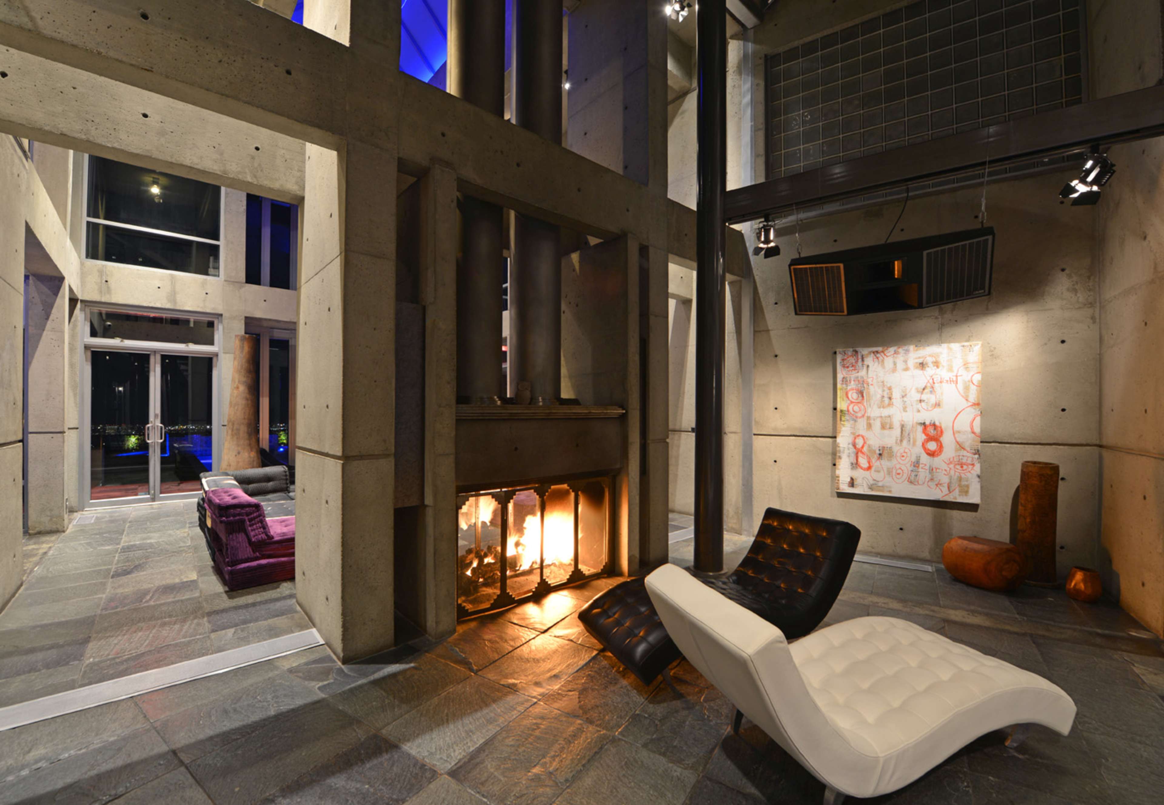The image shows a modern interior space with concrete walls, a fireplace, a white chaise lounge, and a colorful artwork on display.