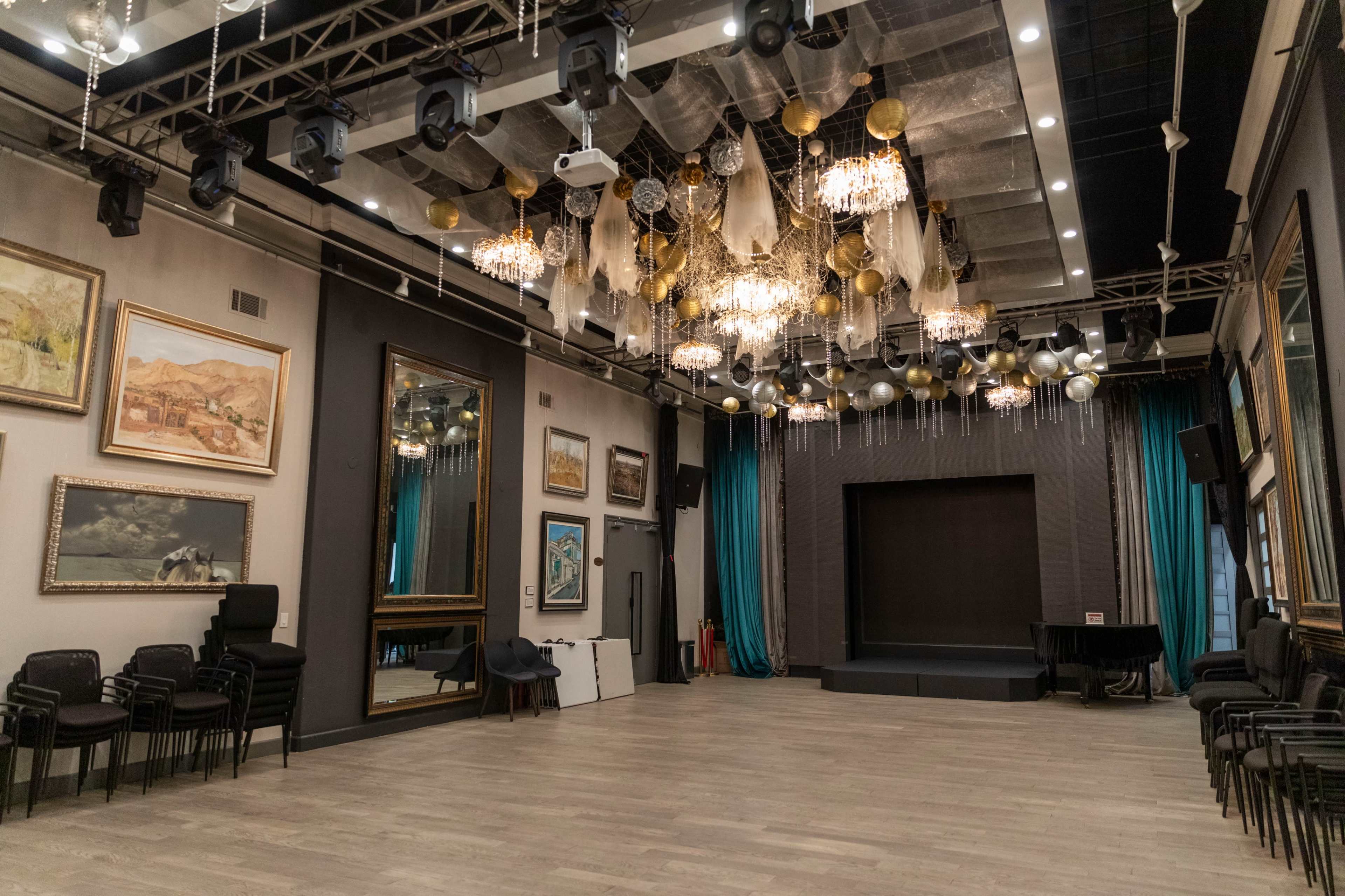 The image shows an empty event hall featuring a stage, decorative chandeliers, and various artworks on the walls.