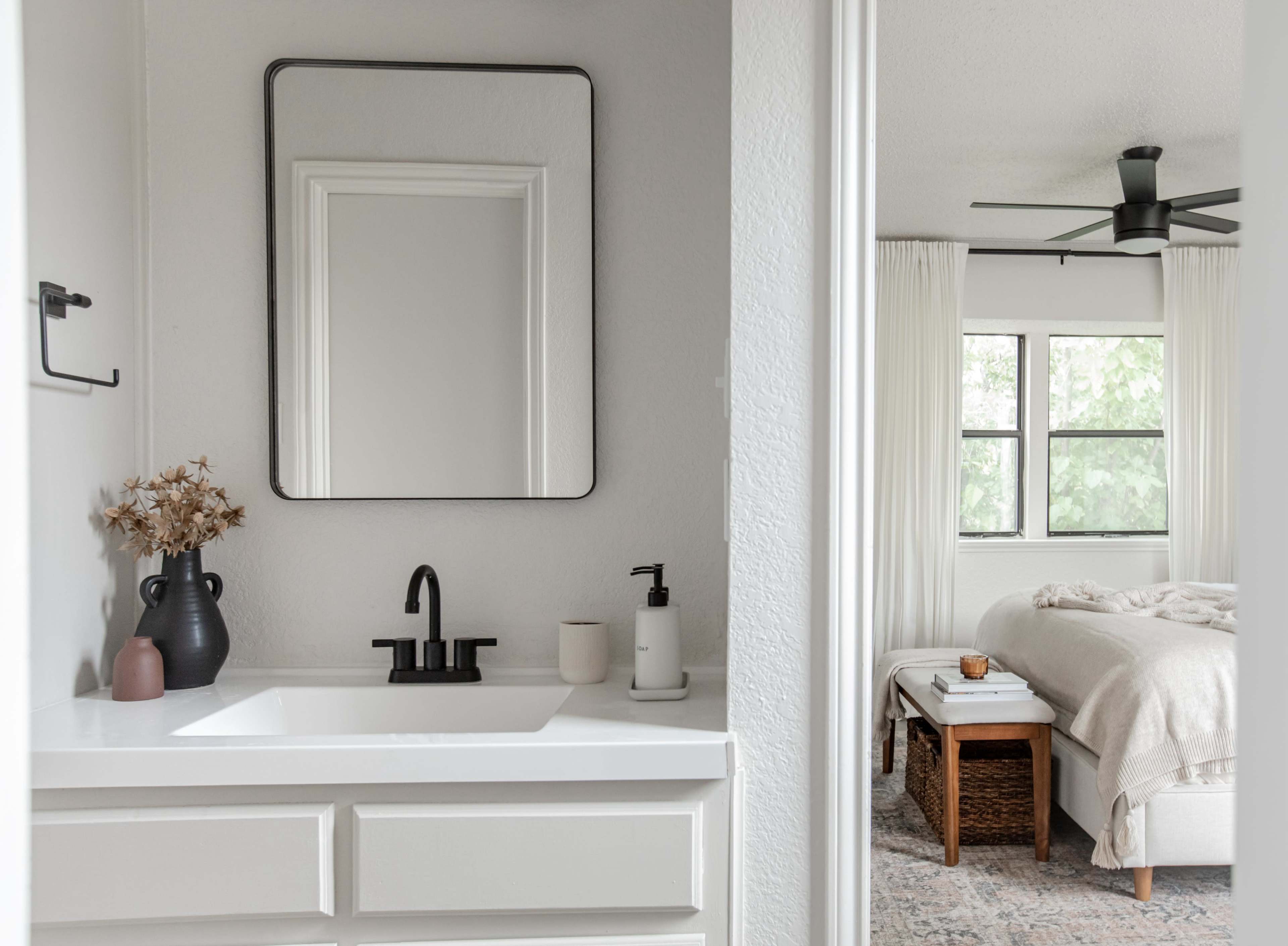 A modern bathroom with a white sink and a mirror, leading into a bedroom featuring a bed with light bedding and a wicker storage unit.