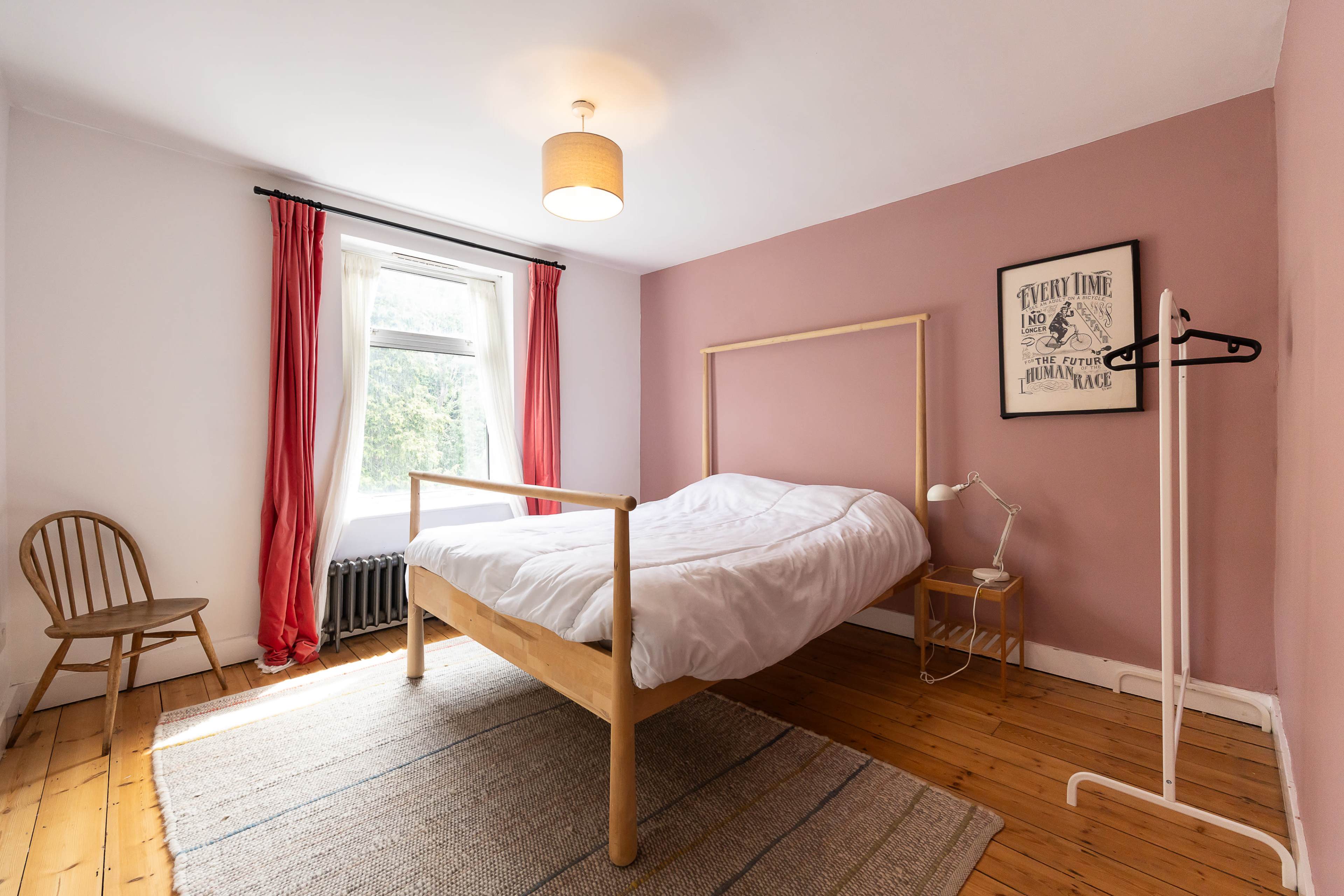 The image shows a simple bedroom featuring a wooden bed frame with a white bedspread, a chair, a floor lamp, and a window with sheer curtains.