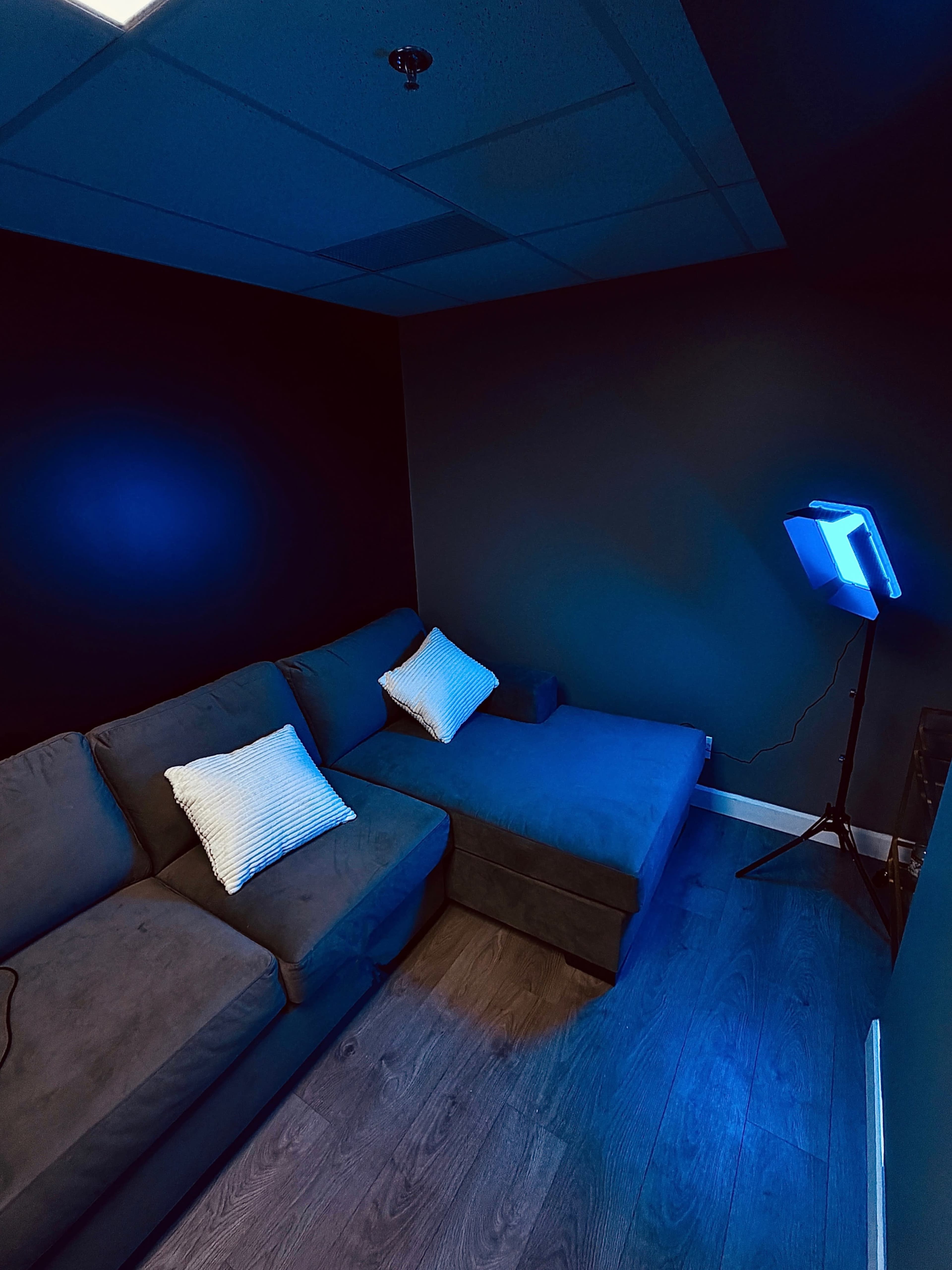 The image shows a small lounge area featuring a dark-colored sectional sofa with two decorative pillows and a floor lamp emitting blue light in a room with dark walls.