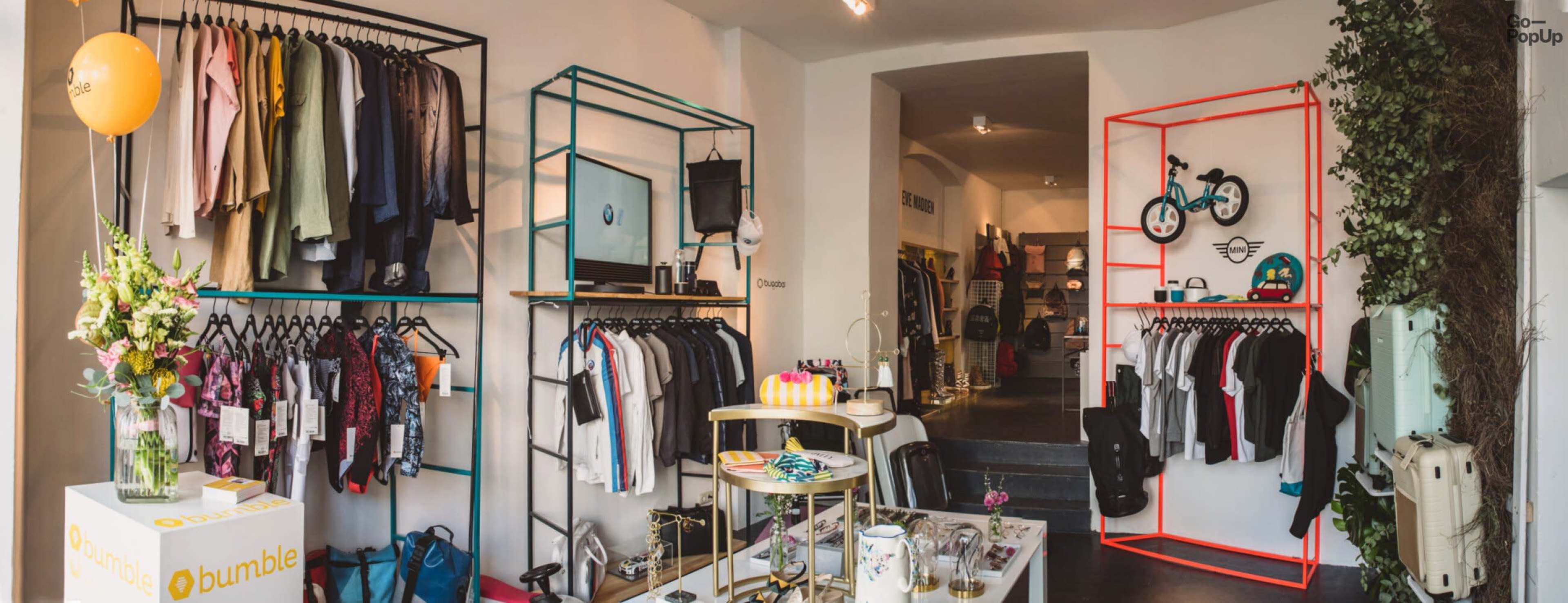 A modern boutique interior featuring clothing racks in various colors, with some clothing items displayed alongside accessories and decor items on a table.