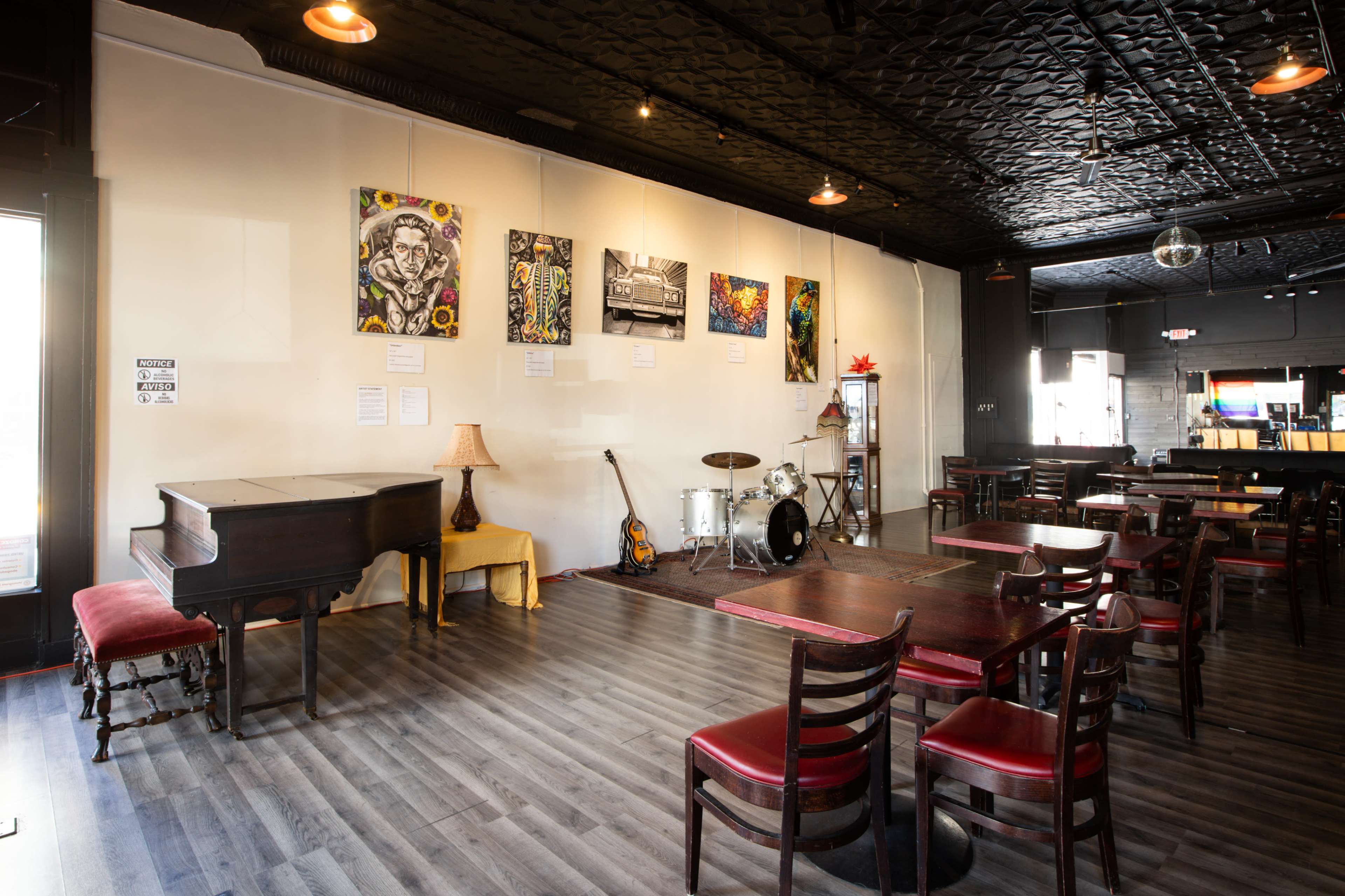 The image shows a spacious café or performance venue with a piano, a small stage area with a drum set, and colorful artwork displayed on the walls.