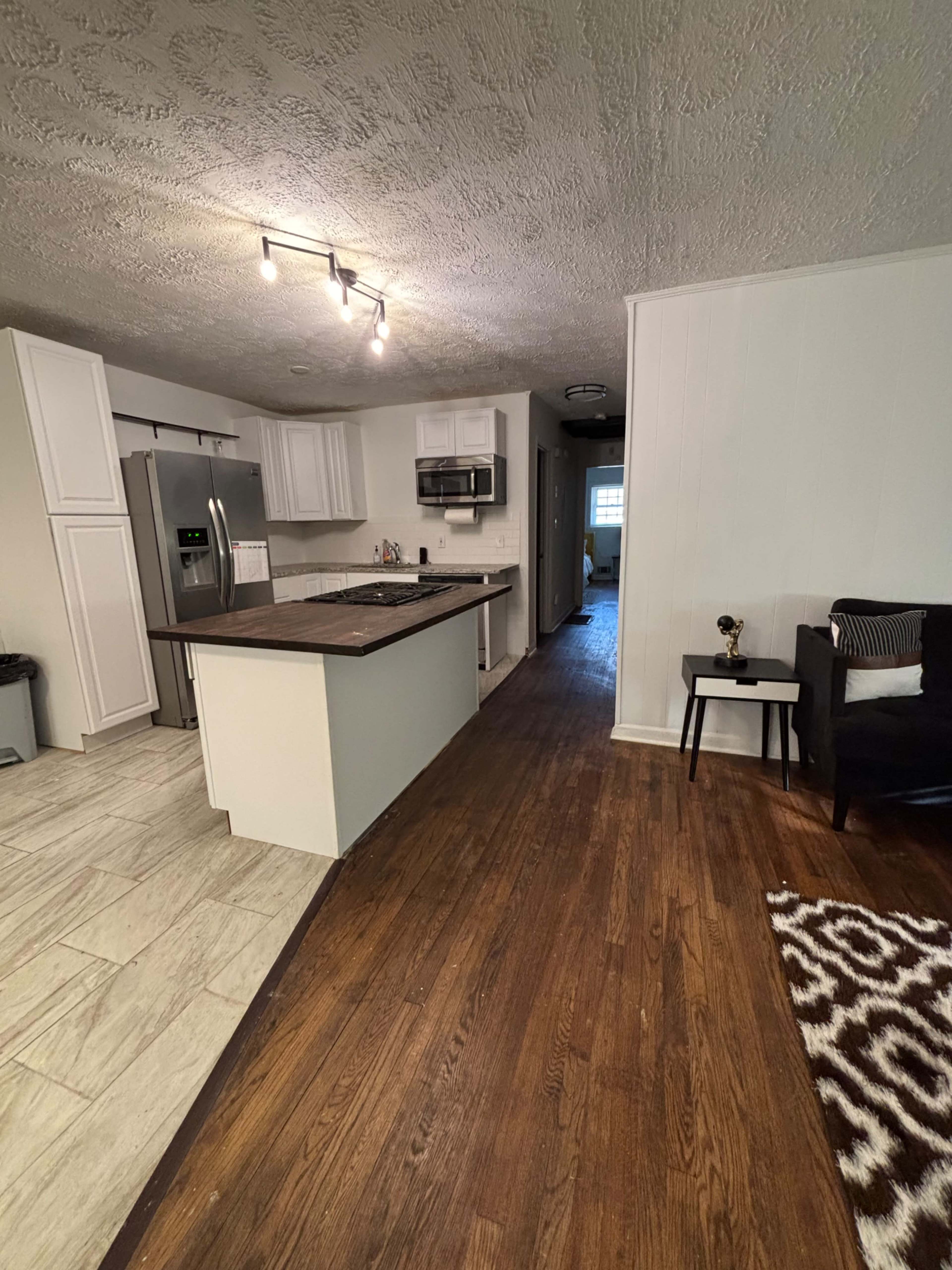 The image shows a kitchen and living area with white cabinets, stainless steel appliances, and hardwood flooring leading to a hallway.