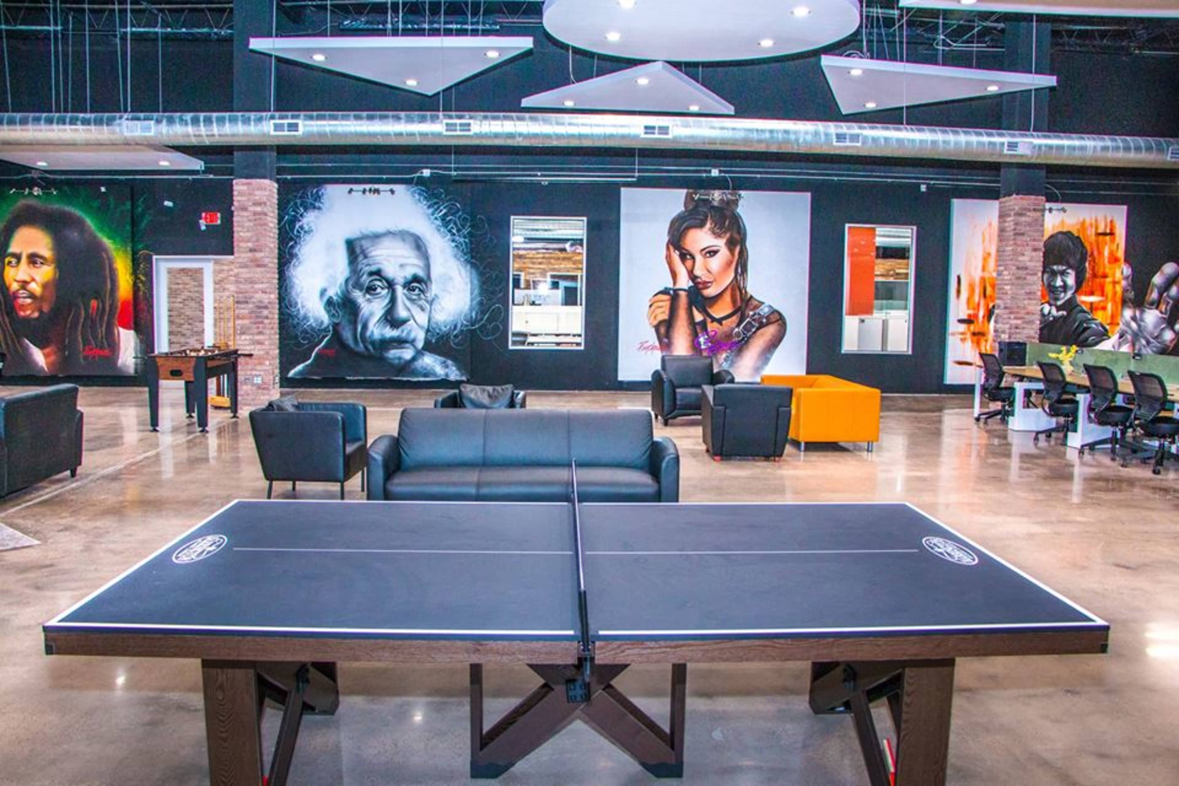 A modern lounge area featuring a ping pong table in the foreground, surrounded by large wall murals of famous figures and comfortable seating arrangements.