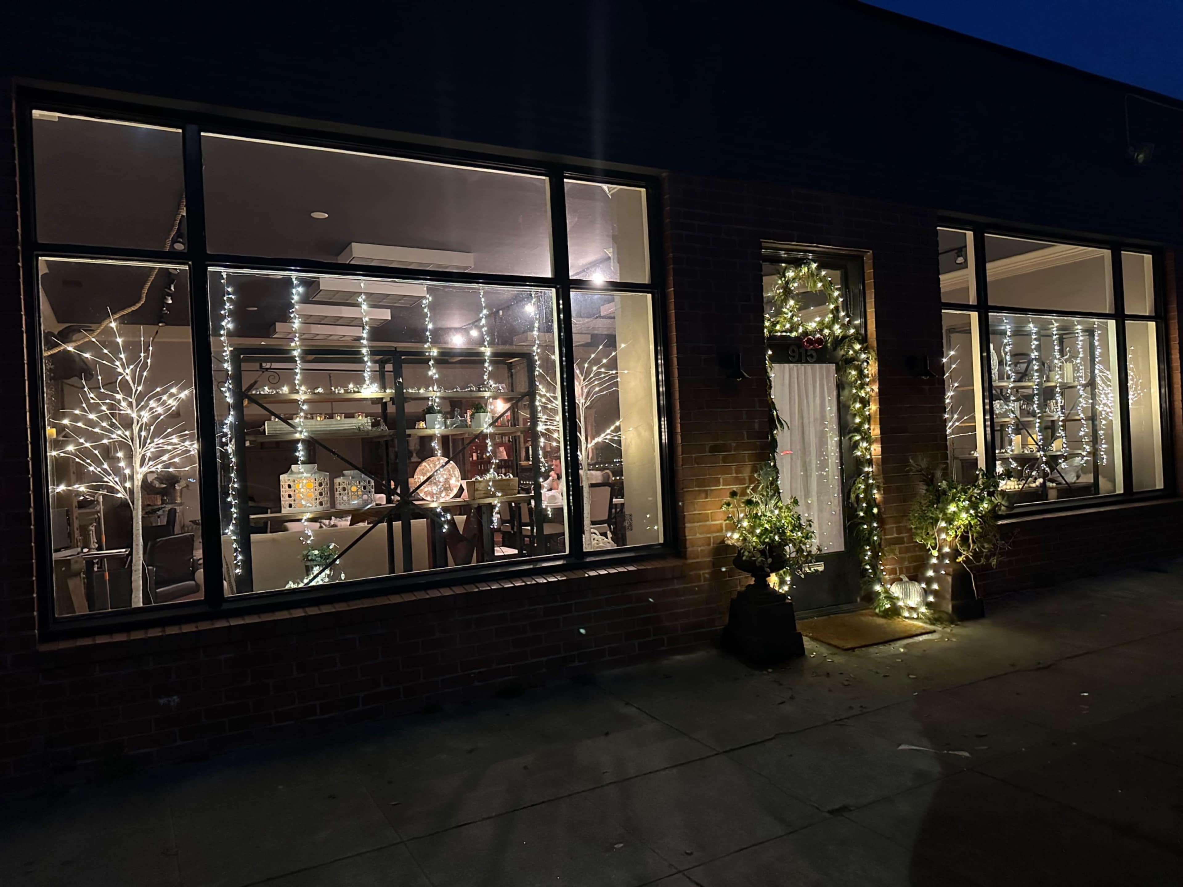 The storefront features large windows illuminated with string lights and decorated with small trees, showcasing a cozy interior filled with shelves and products.
