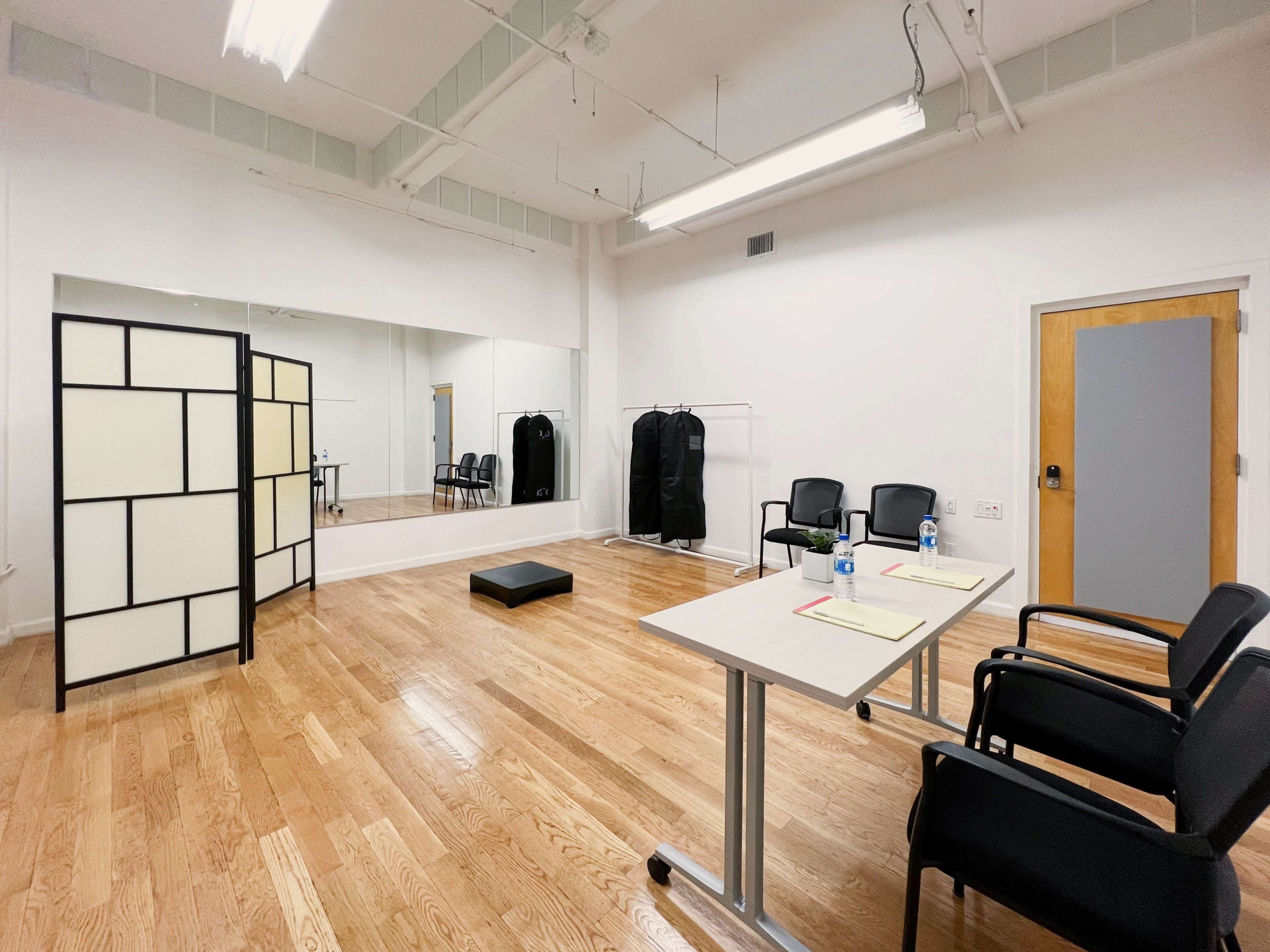 The image shows a spacious, empty room with light wood flooring, two tables with chairs, a partition, and a large mirror.