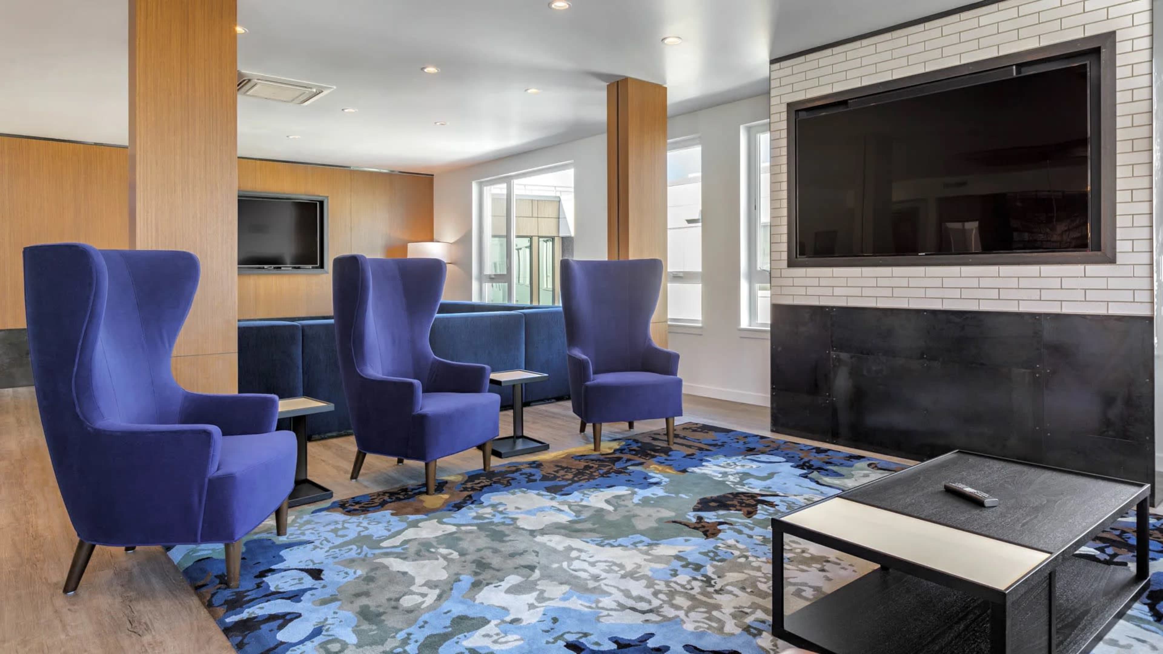 A modern lounge area featuring two blue armchairs, a patterned rug, and a flat-screen television mounted on a white brick wall.