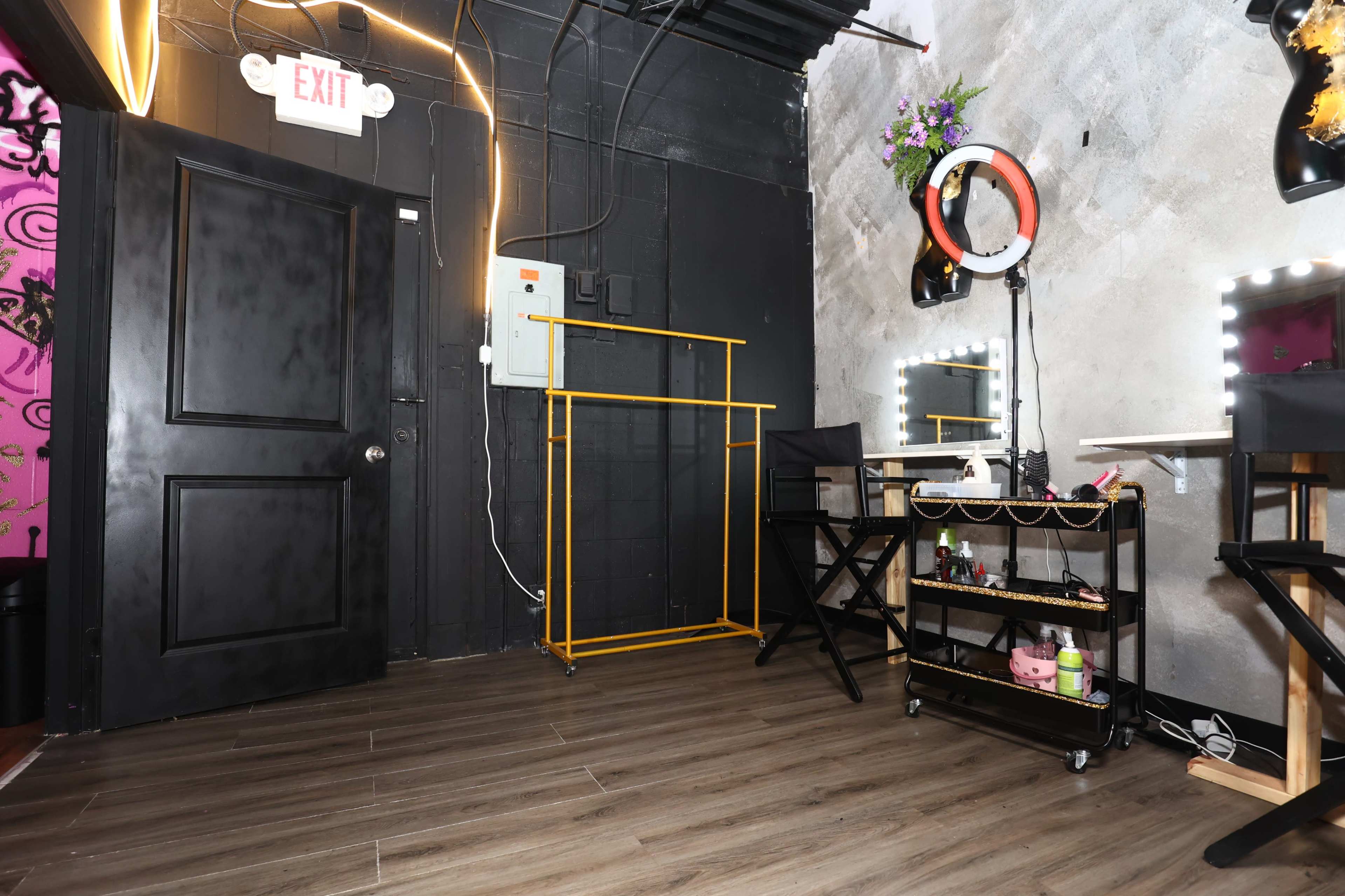 The image shows a small, dimly lit room featuring a black door, a clothing rack in yellow, a mirror with lights, and a black cart with various items.