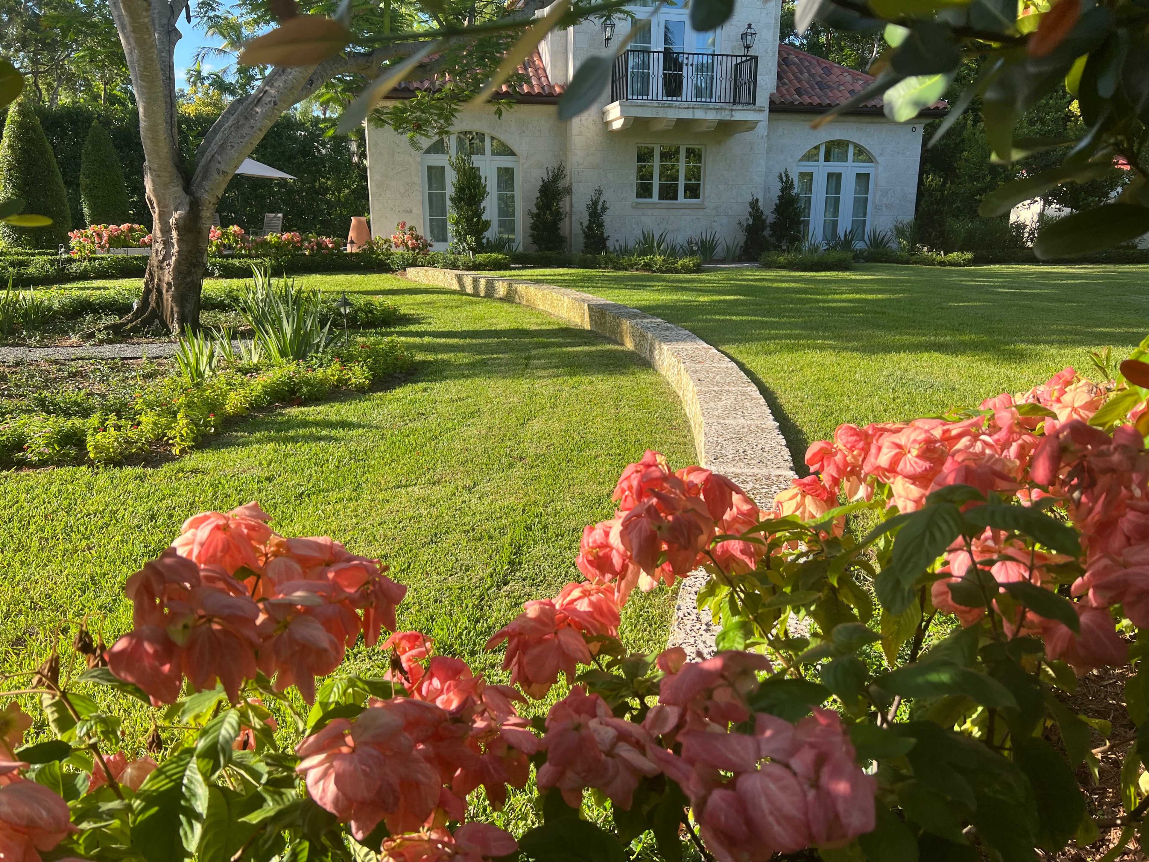 A curved stone path leads through a manicured lawn to a house surrounded by landscaped gardens and blooming pink flowers.
