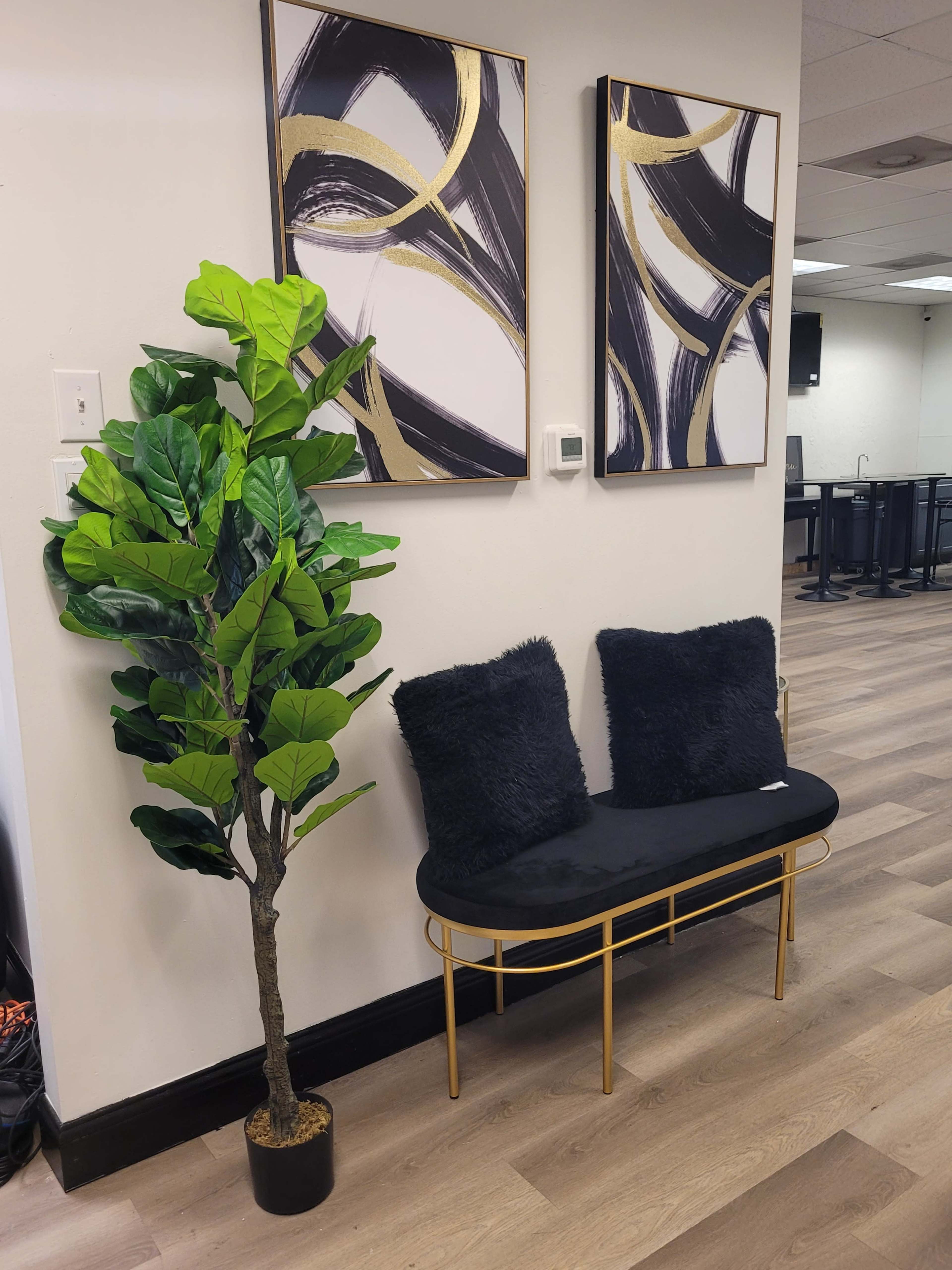 A small seating area features a gold-framed bench with black cushions, complemented by a potted leafy plant and two abstract art pieces on the wall.