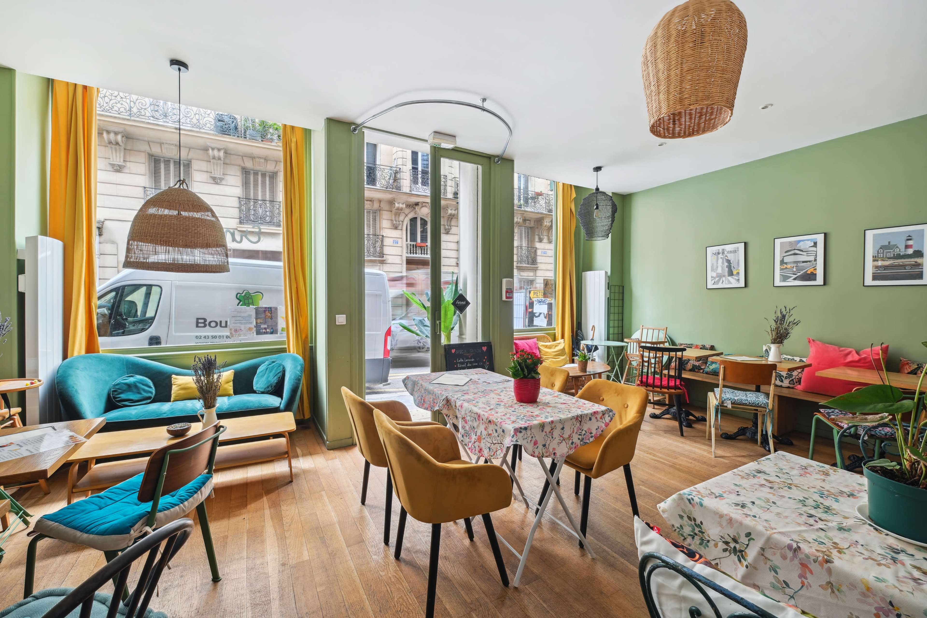 A cozy café features colorful seating, floral tablecloths, and large windows allowing natural light to fill the space.