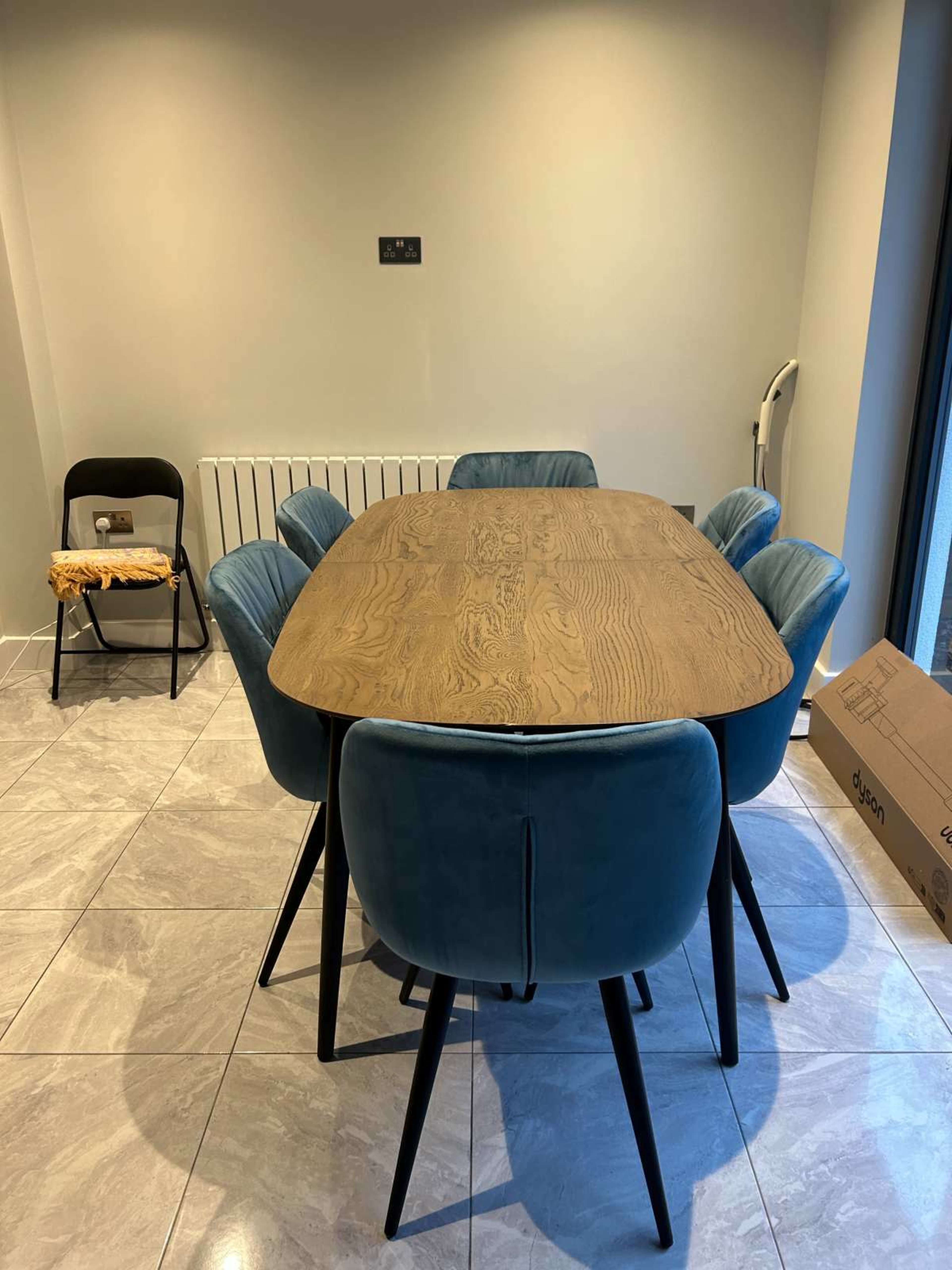 A wooden dining table surrounded by six blue upholstered chairs is placed in a room with tiled flooring.