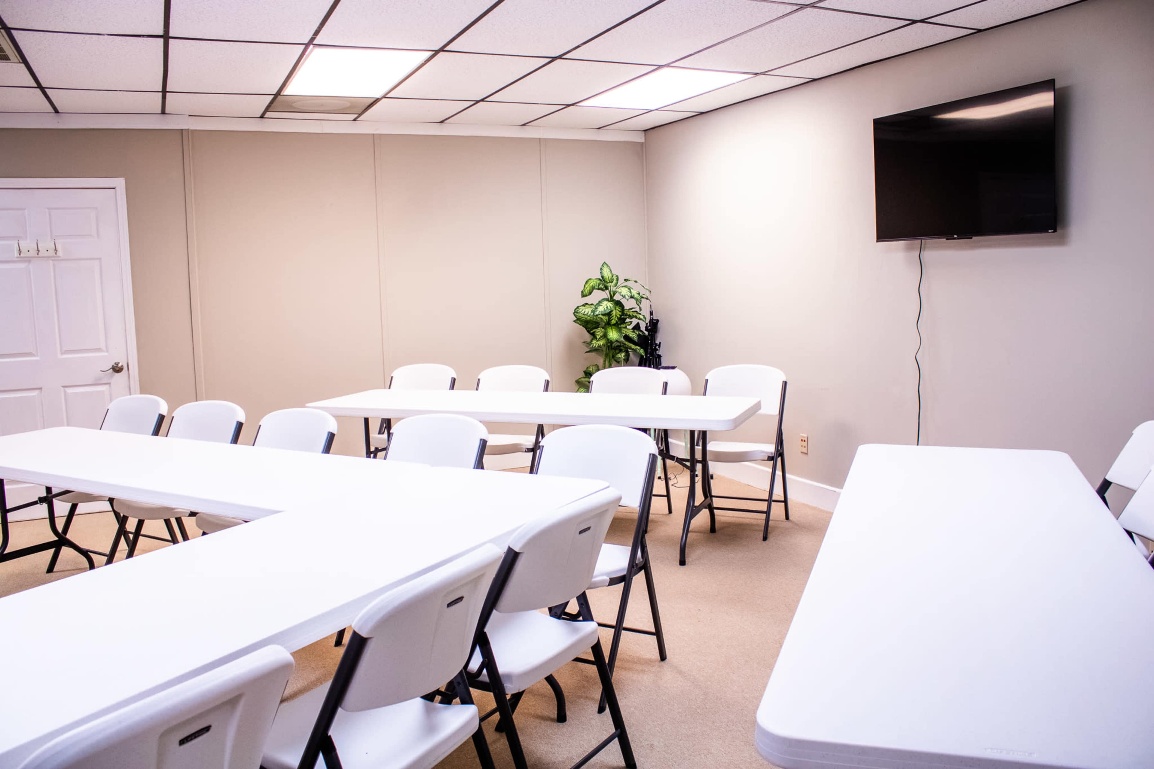 The image shows a meeting room with several white tables and chairs arranged around the space, and a television mounted on the wall.