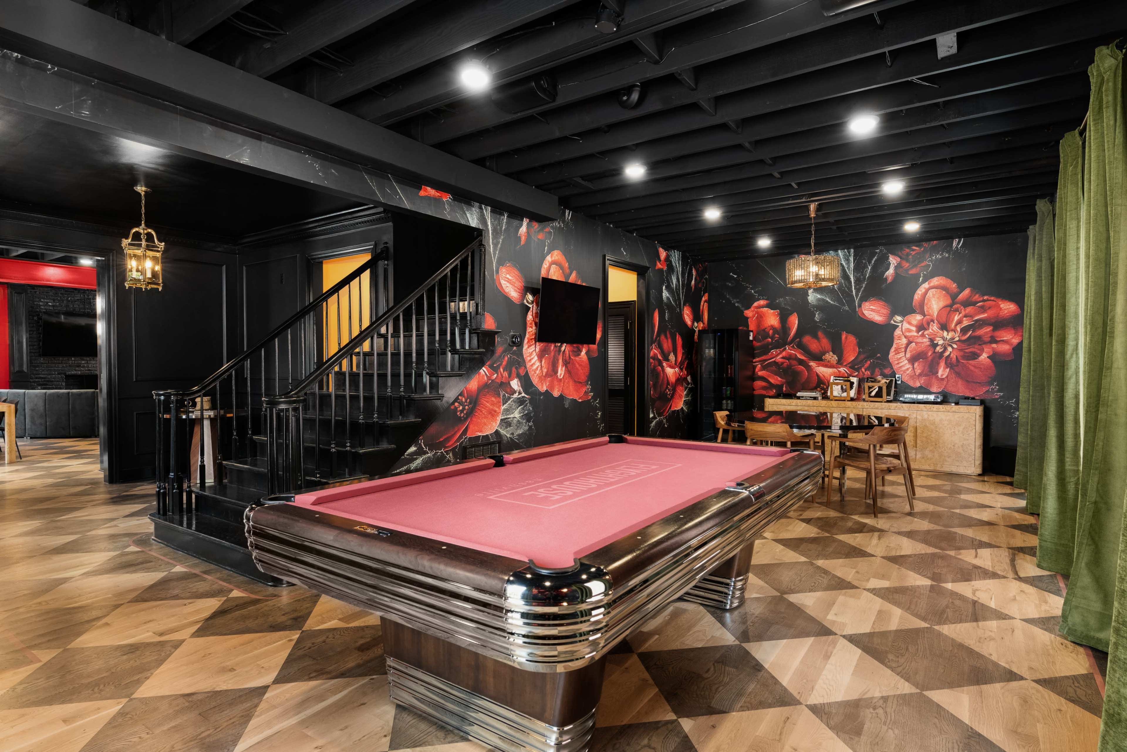 A stylish game room featuring a pink pool table, dark floral wallpaper, and a dining area with wooden furniture.